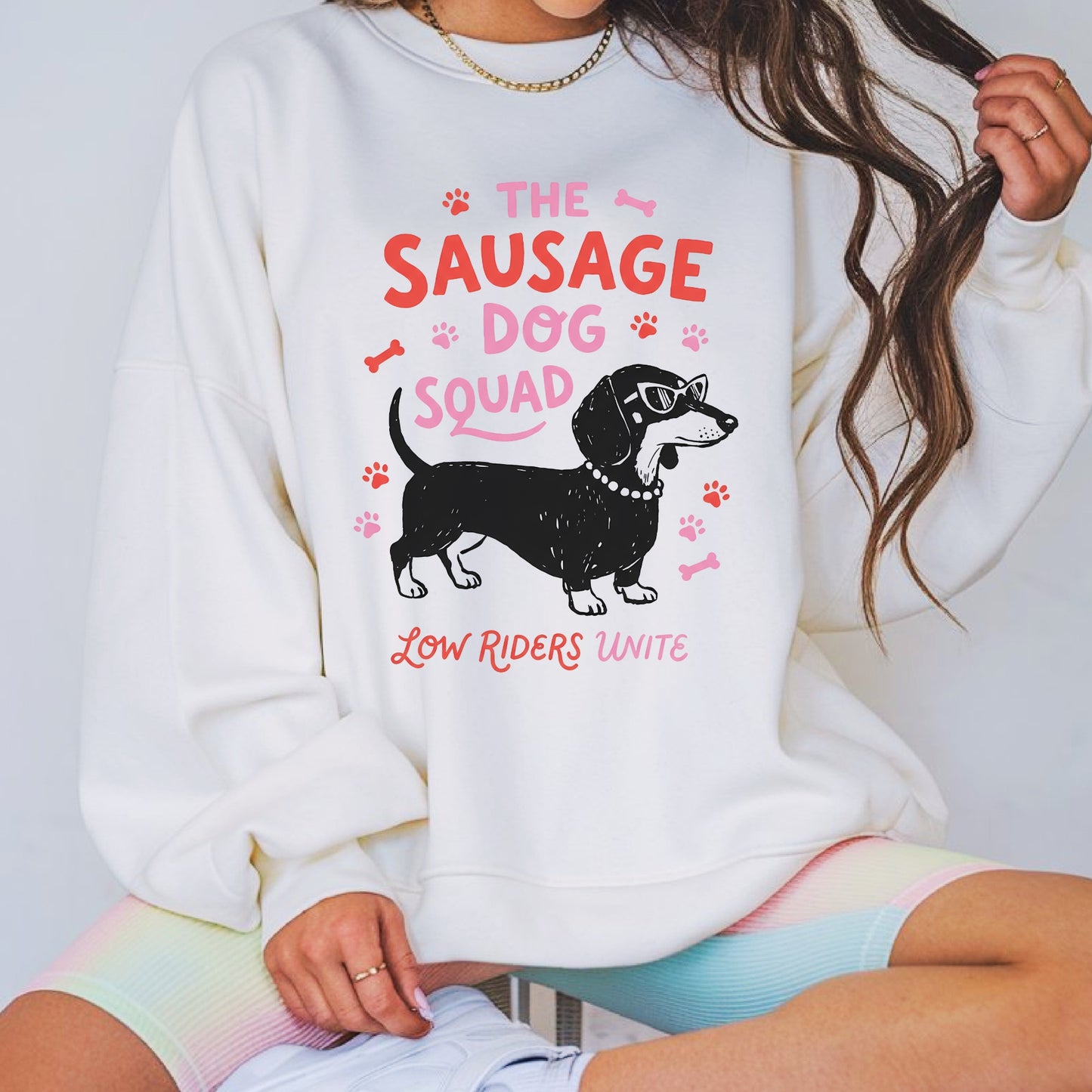 Funny dachshund graphic featuring a stylish sausage dog wearing sunglasses with the phrase The Sausage Dog Squad Low Riders Unite, surrounded by paw prints and bones in pink and coral tones