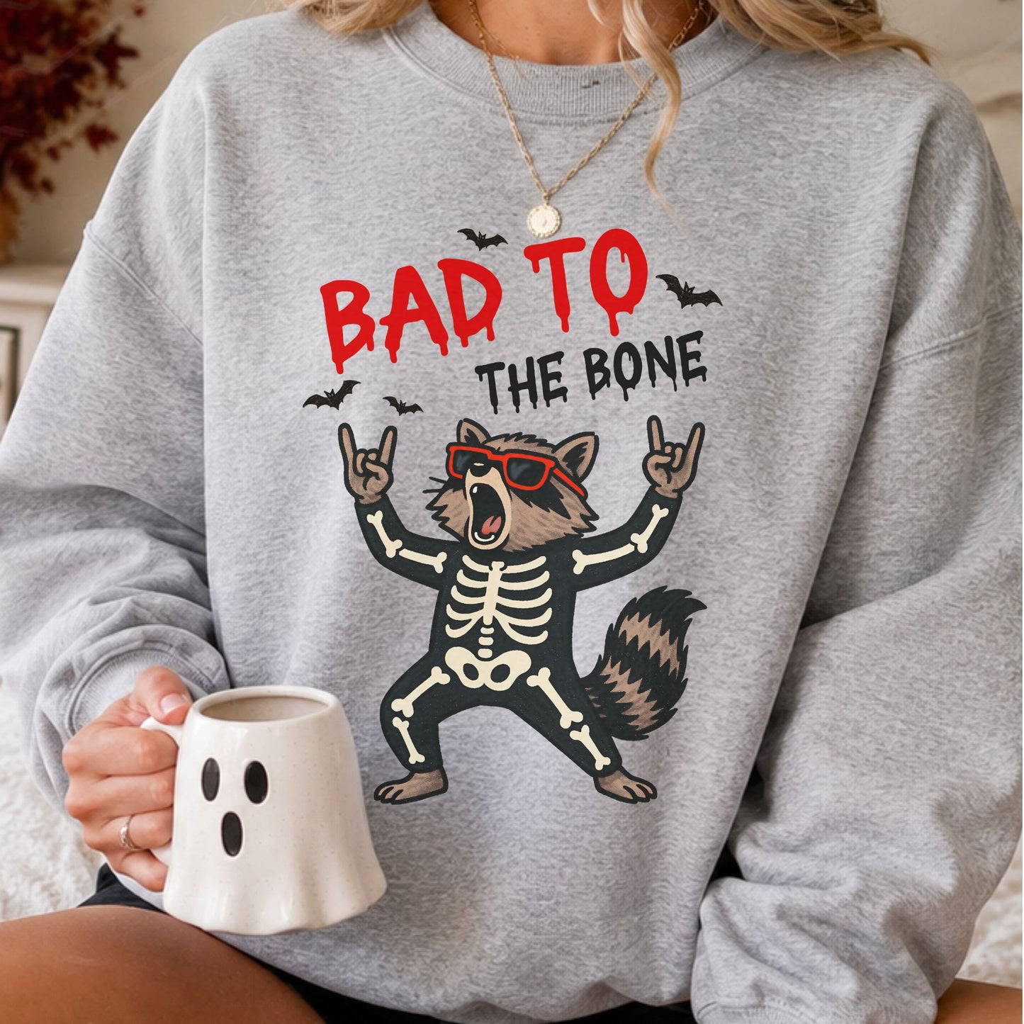 Bad to the Bone Halloween DTF transfer featuring a funny raccoon wearing a skeleton costume and red sunglasses, surrounded by black bats on a gray sweatshirt mockup.