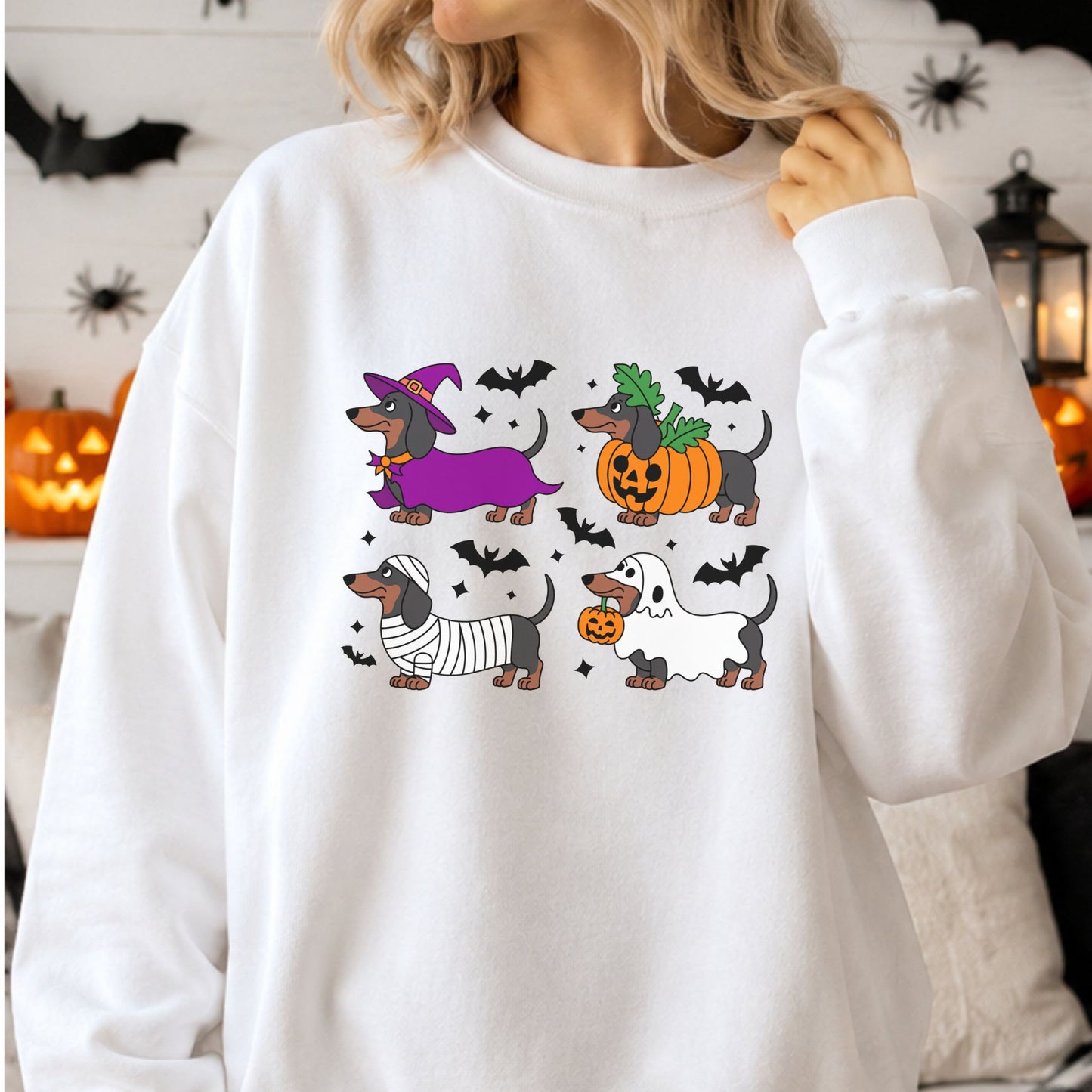 Cute Halloween dachshund DTF transfer featuring four wiener dogs dressed in witch, jack-o-lantern pumpkin, mummy and ghost costumes surrounded by black bats on a white sweatshirt mockup.