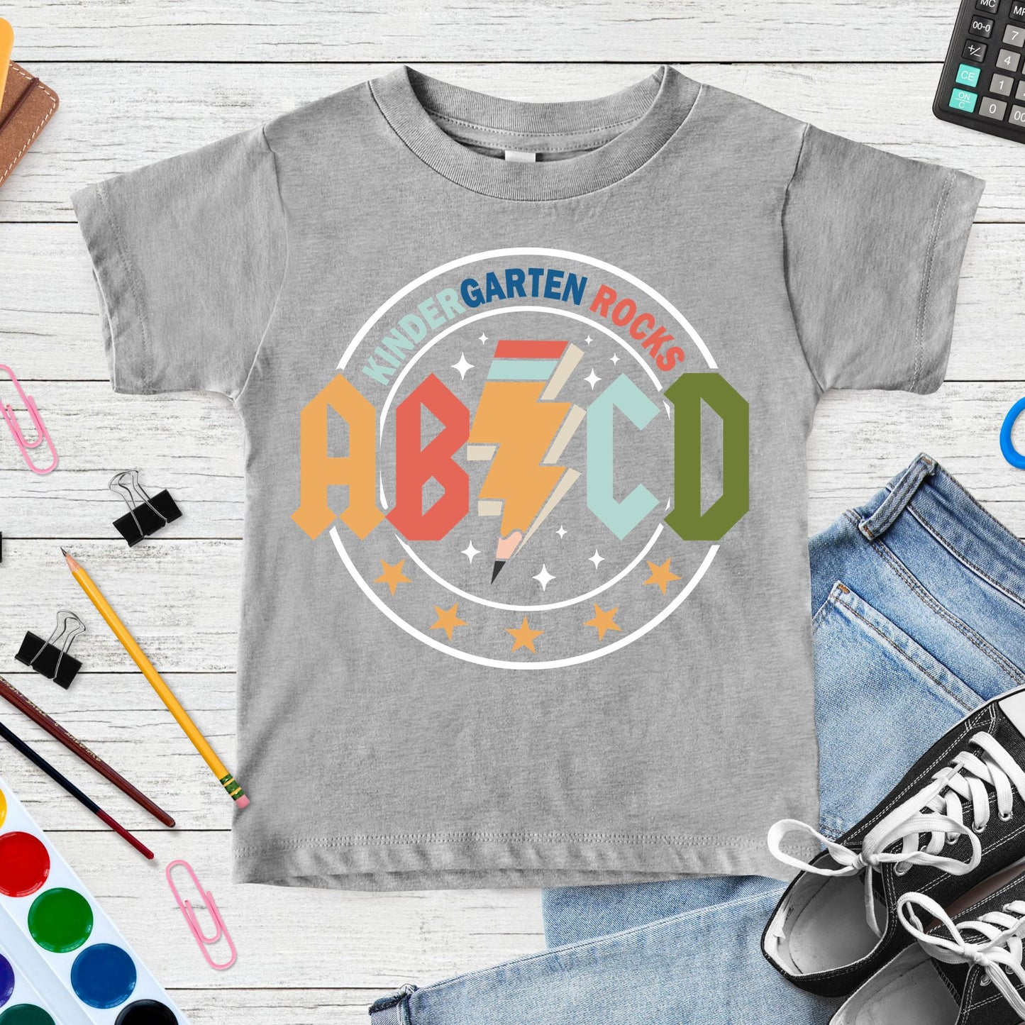 Kindergarten Rocks back-to-school design featuring bold ABCD letters, a pencil-shaped lightning bolt, stars, sparkles, and colorful retro school graphics for kids shirts.