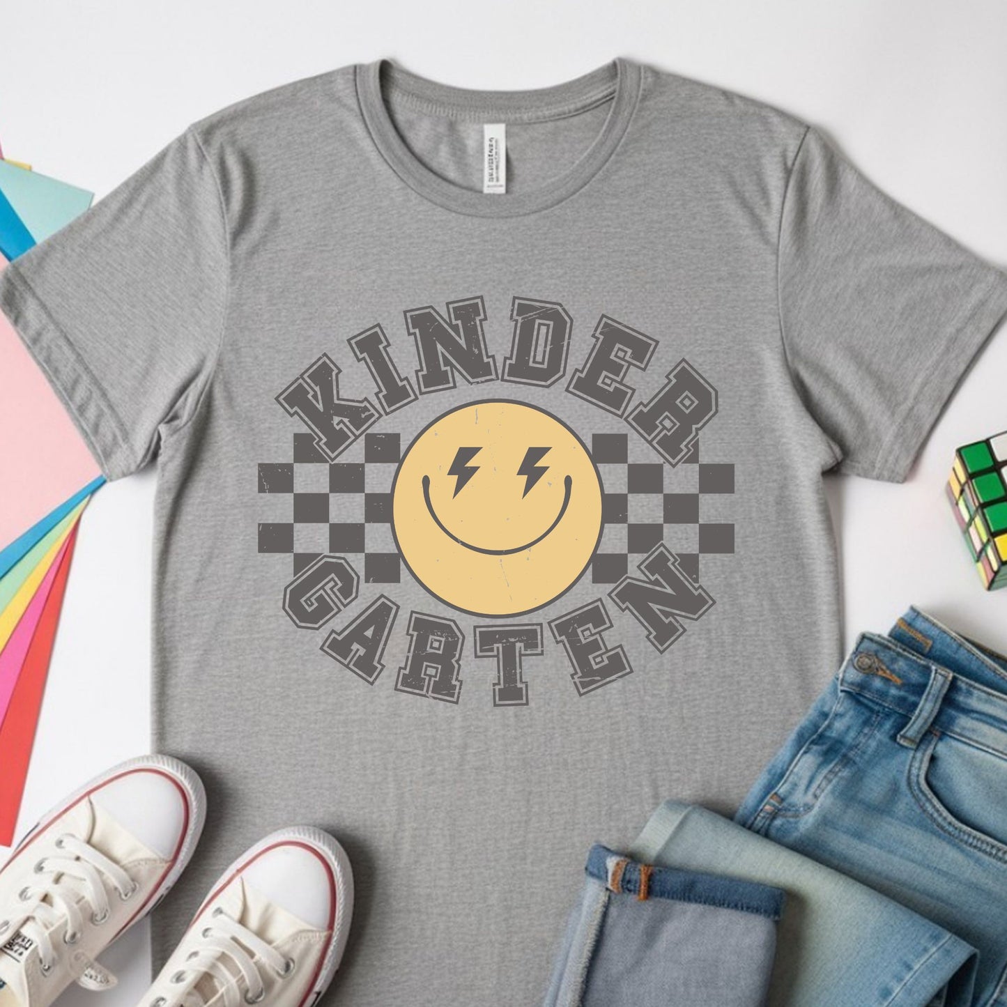 Kindergarten back-to-school design featuring distressed collegiate lettering, a yellow smiley face with lightning-bolt eyes, and bold black checkerboard accents for kids shirts.