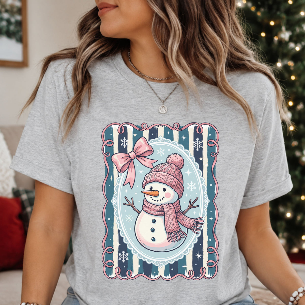 Snowman wearing a pink hat and scarf with a faux knit pattern, surrounded by a pale blue oval frame, navy and teal stripes, a pink bow and white snowflakes.