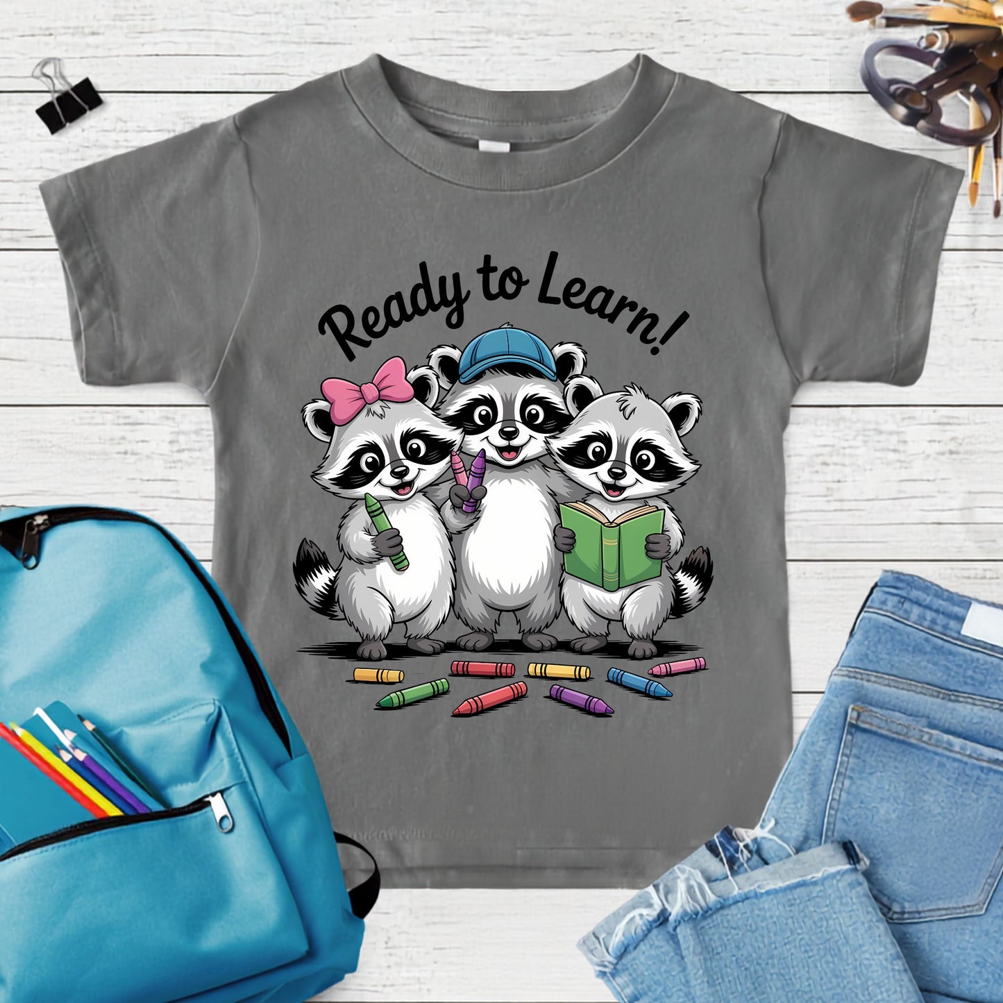 Ready to Learn back-to-school design featuring three cute raccoons holding crayons and a book, with colorful school supplies and playful classroom details.