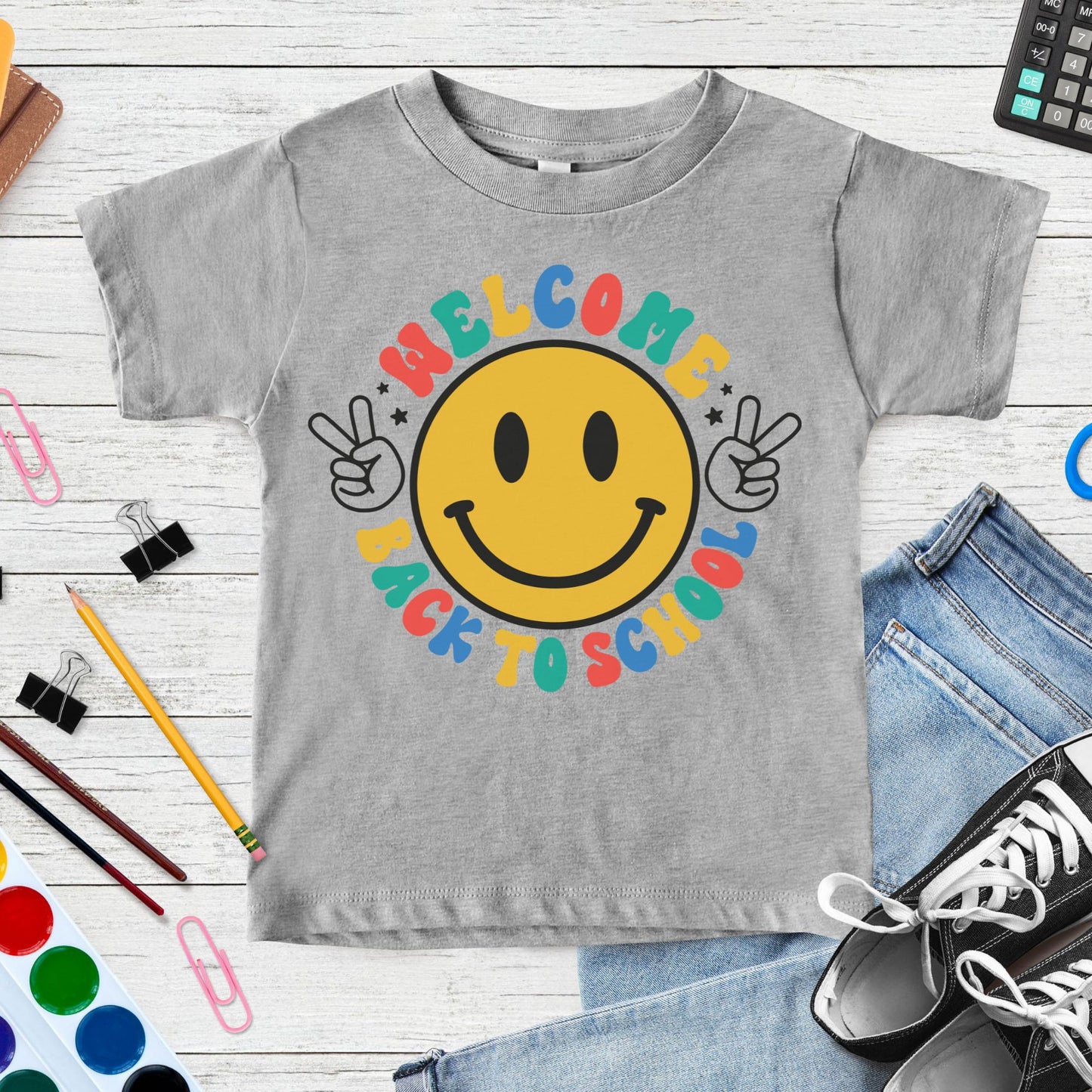 Welcome Back to School design featuring a large yellow smiley face, peace-sign hands, colorful retro lettering, and black star accents for toddler, kids, and youth shirts.