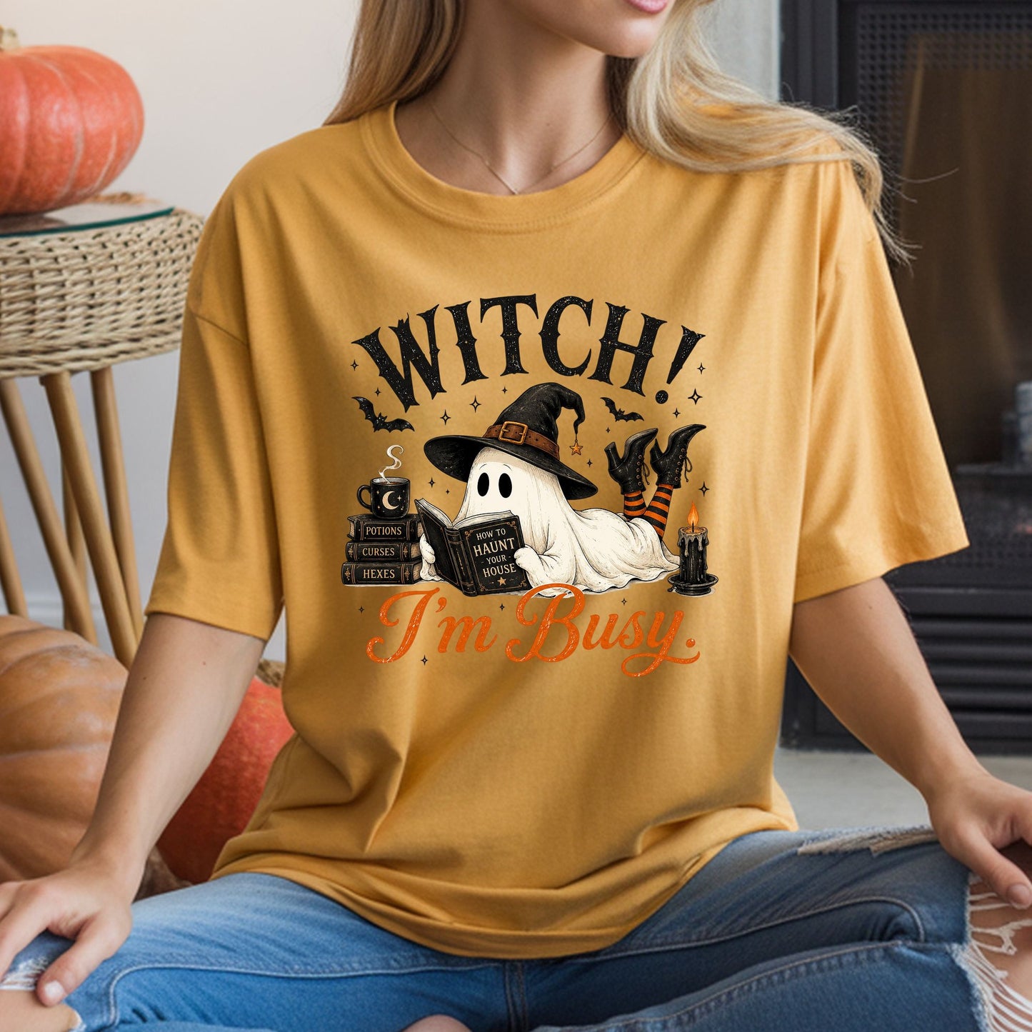 Witch I’m Busy Halloween DTF transfer featuring a sheet ghost wearing a witch hat and striped stockings while lying down reading a How to Haunt Your House book, surrounded by potion and spell books, a steaming moon mug, candle, bats and sparkling stars on a mustard yellow T-shirt mockup.