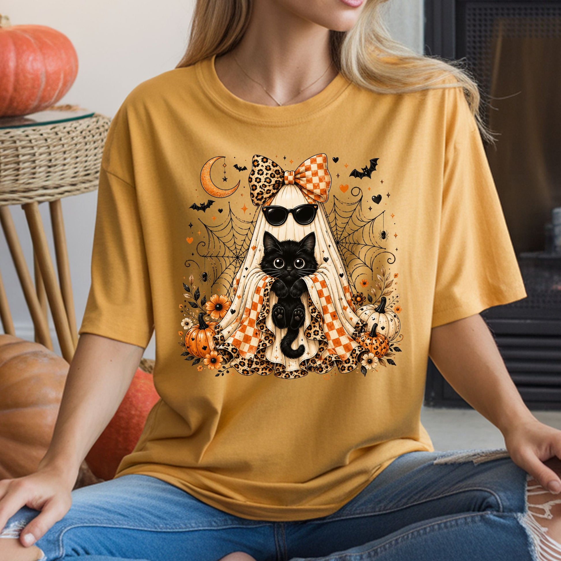 Coquette Halloween DTF transfer featuring a stylish sheet ghost holding a cute black cat, wearing sunglasses and an oversized leopard print and orange gingham bow, surrounded by pumpkins, flowers, spiderwebs, bats and a crescent moon on a mustard yellow T-shirt mockup.
