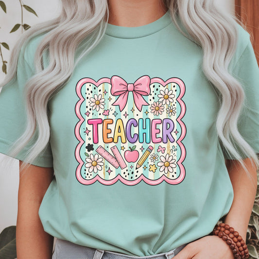Cute coquette teacher graphic featuring a pink bow, scalloped frame, daisies, apple, ruler, pencil, and pastel classroom elements for teacher shirts, hoodies, and back-to-school apparel.

