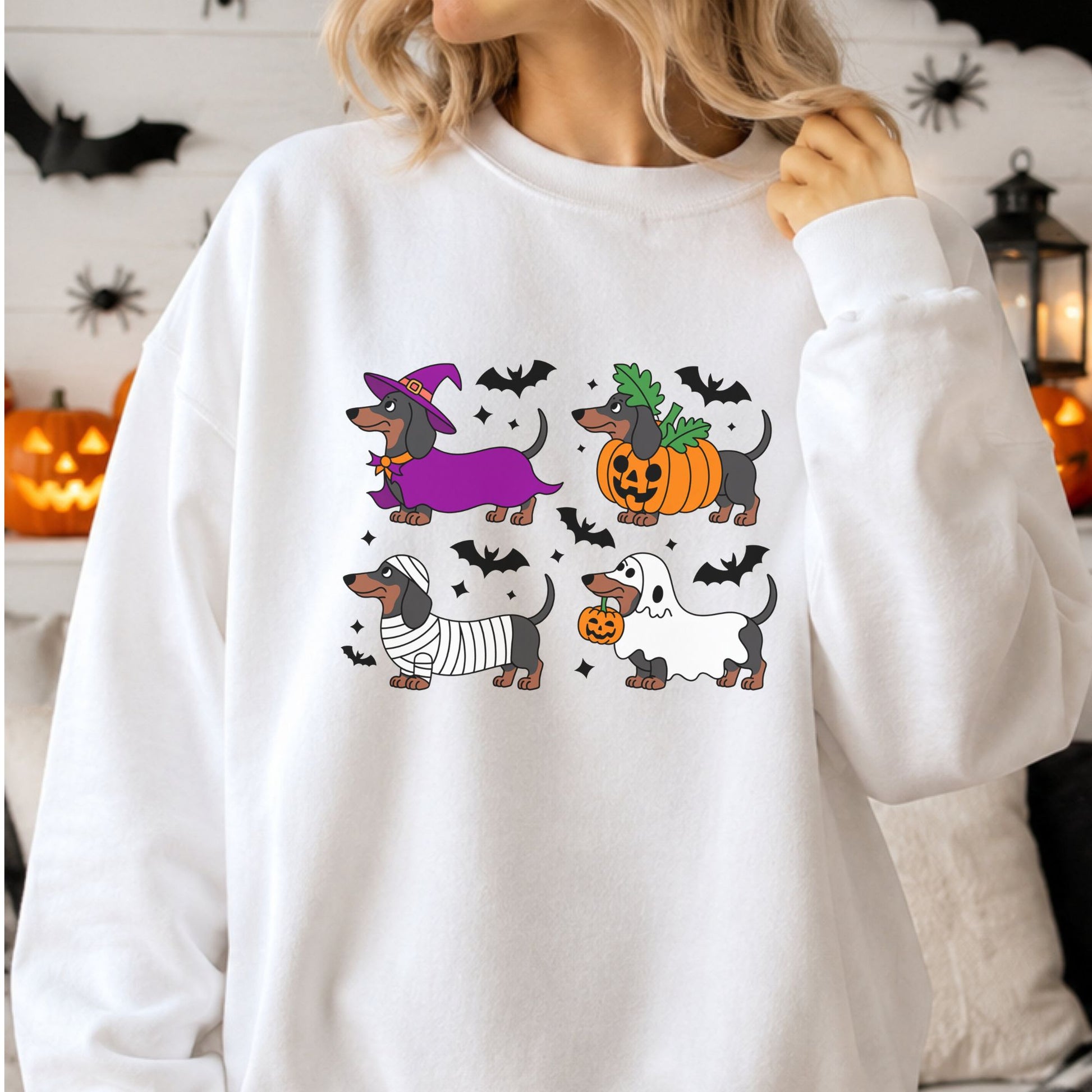 Cute Halloween dachshund DTF transfer featuring four wiener dogs dressed in witch, jack-o-lantern pumpkin, mummy and ghost costumes surrounded by black bats on a white sweatshirt mockup.