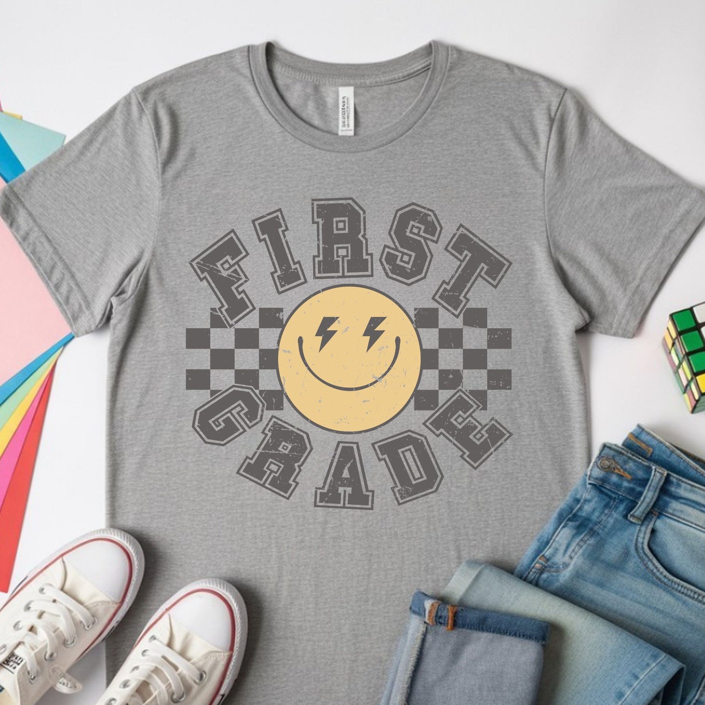 First Grade back-to-school design featuring distressed collegiate lettering, a yellow smiley face with lightning-bolt eyes, and bold black checkerboard accents for kids shirts.