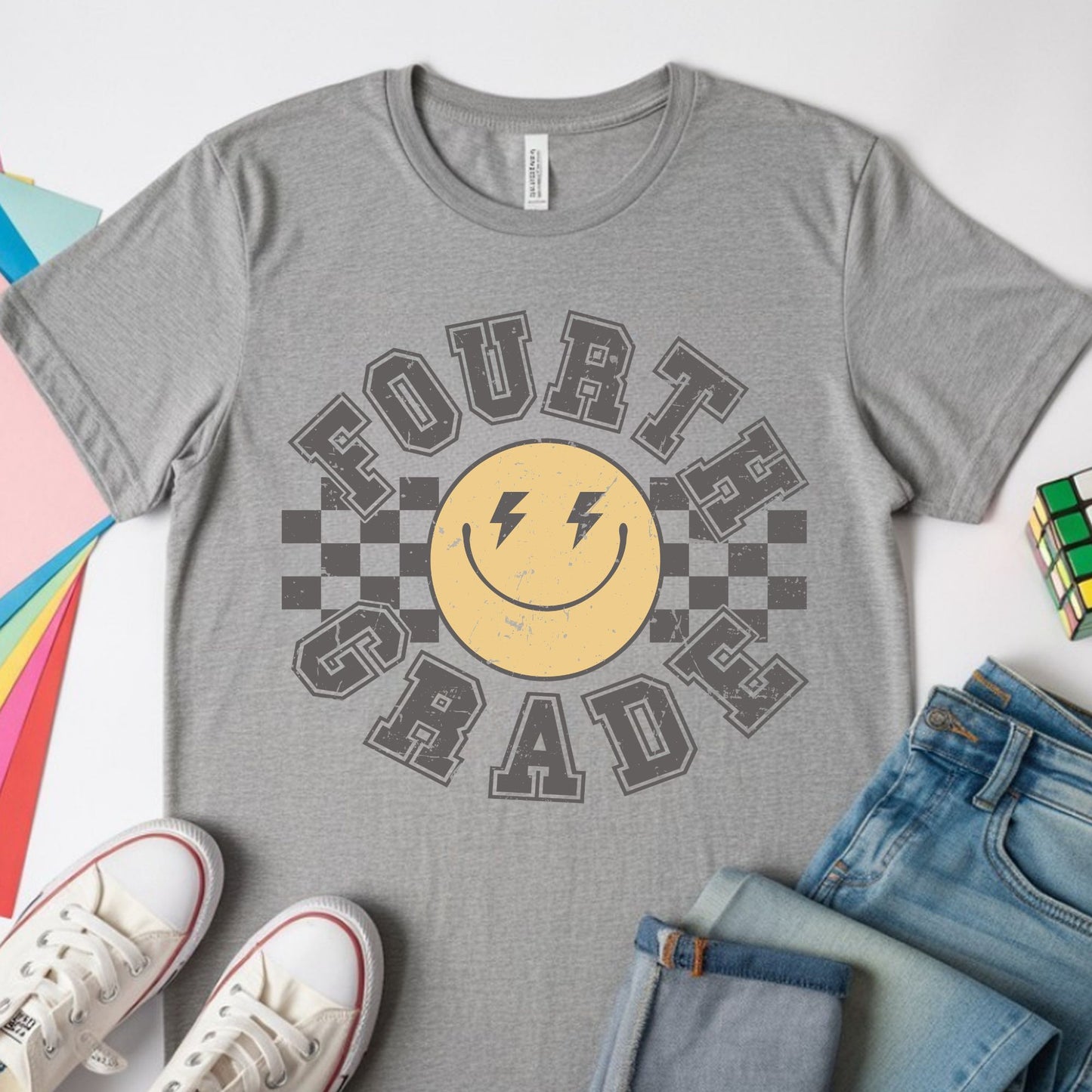 Fourth Grade back-to-school design featuring distressed collegiate lettering, a yellow smiley face with lightning-bolt eyes, and bold black checkerboard accents for kids shirts.