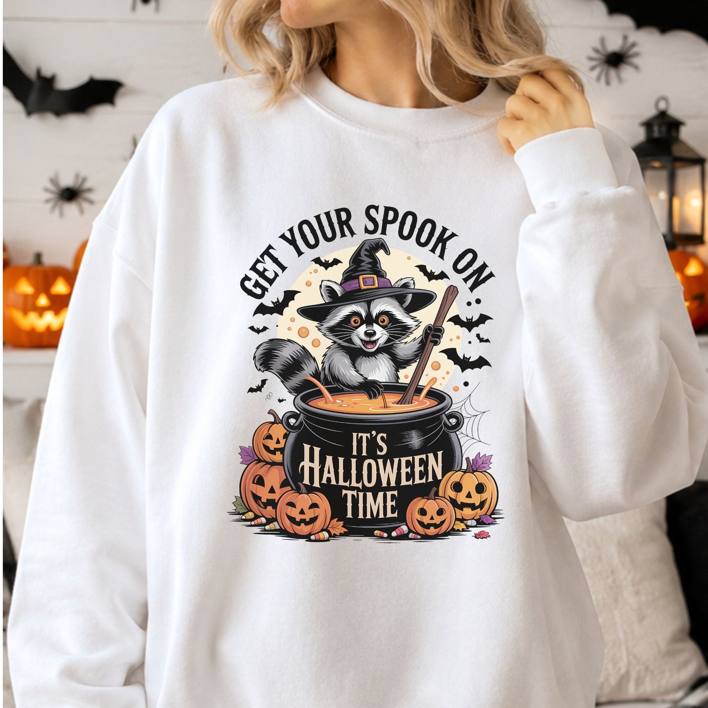 Get Your Spook On Halloween DTF transfer featuring a cheerful raccoon wearing a witch hat and stirring an orange potion in a black cauldron, surrounded by jack-o-lanterns, bats, candy corn, autumn leaves and a spiderweb on a white sweatshirt mockup.