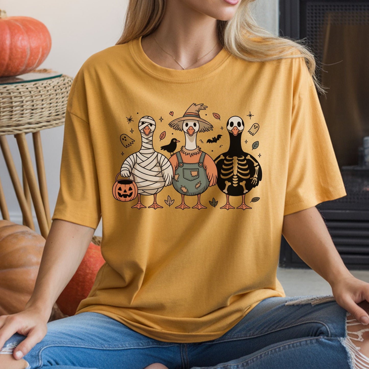 Cute Halloween DTF transfer featuring three geese dressed in mummy, rustic scarecrow and skeleton costumes with a jack-o-lantern candy bucket, bats, tiny ghosts, autumn leaves and a black crow on a mustard yellow T-shirt mockup.