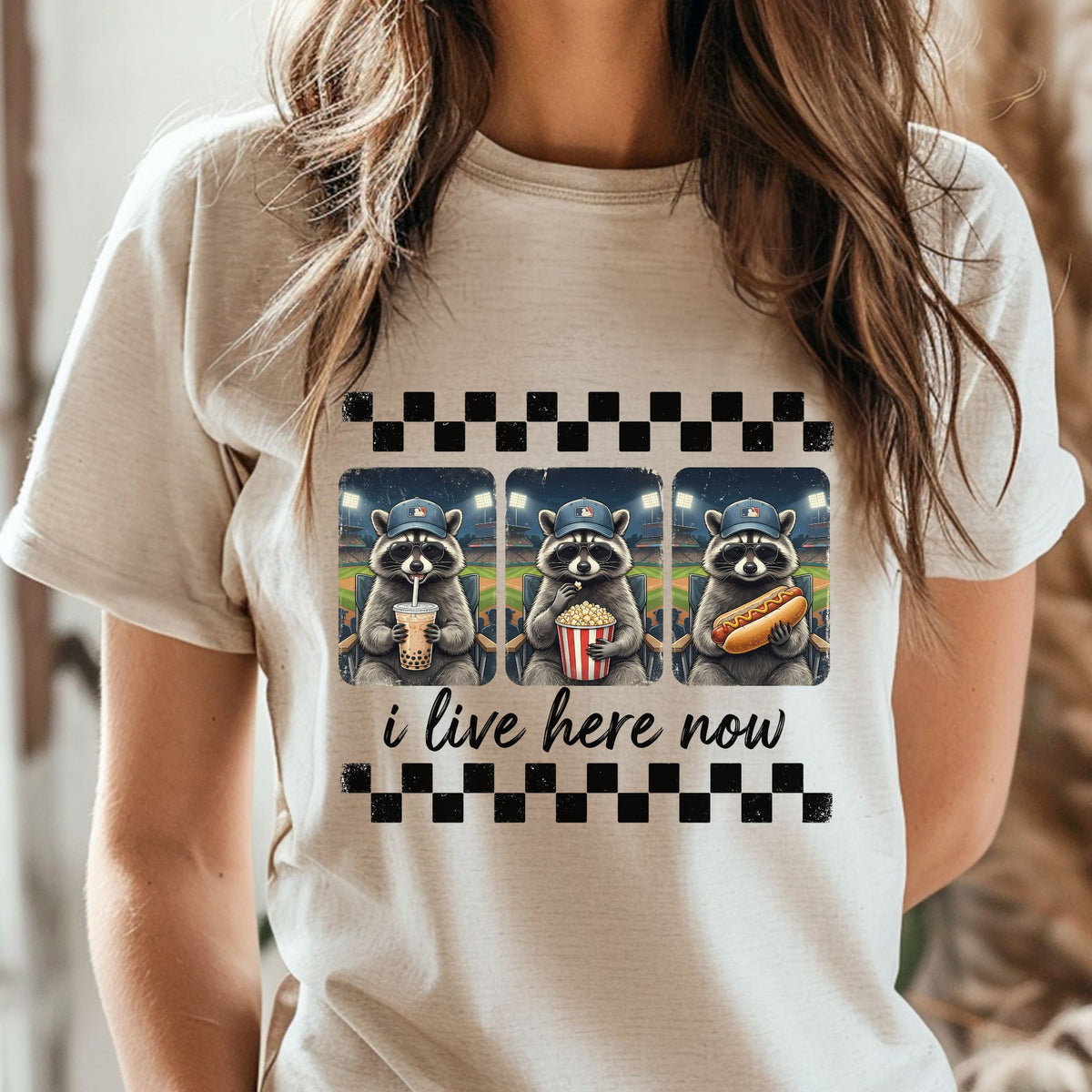 I Live Here Now baseball raccoon DTF transfer featuring three cool raccoons at a baseball game with sunglasses, a baseball cap, boba drink, popcorn, and hot dog, plus distressed checkered accents for funny game day shirts and sweatshirts.