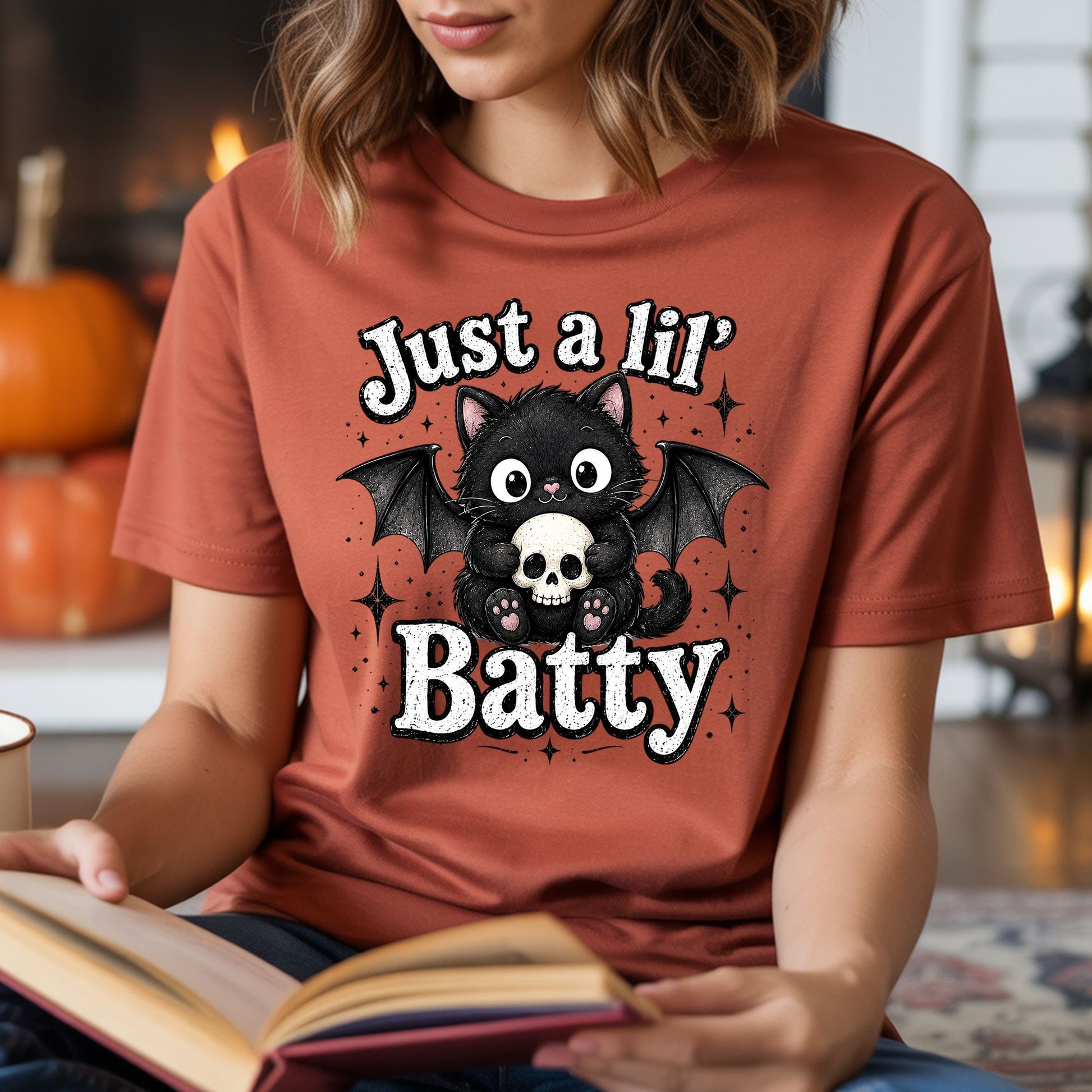 Just a Lil’ Batty Halloween DTF transfer featuring an adorable fluffy black cat with bat wings holding a skull, surrounded by sparkling black stars and bold vintage-style lettering on a rust-colored T-shirt mockup.