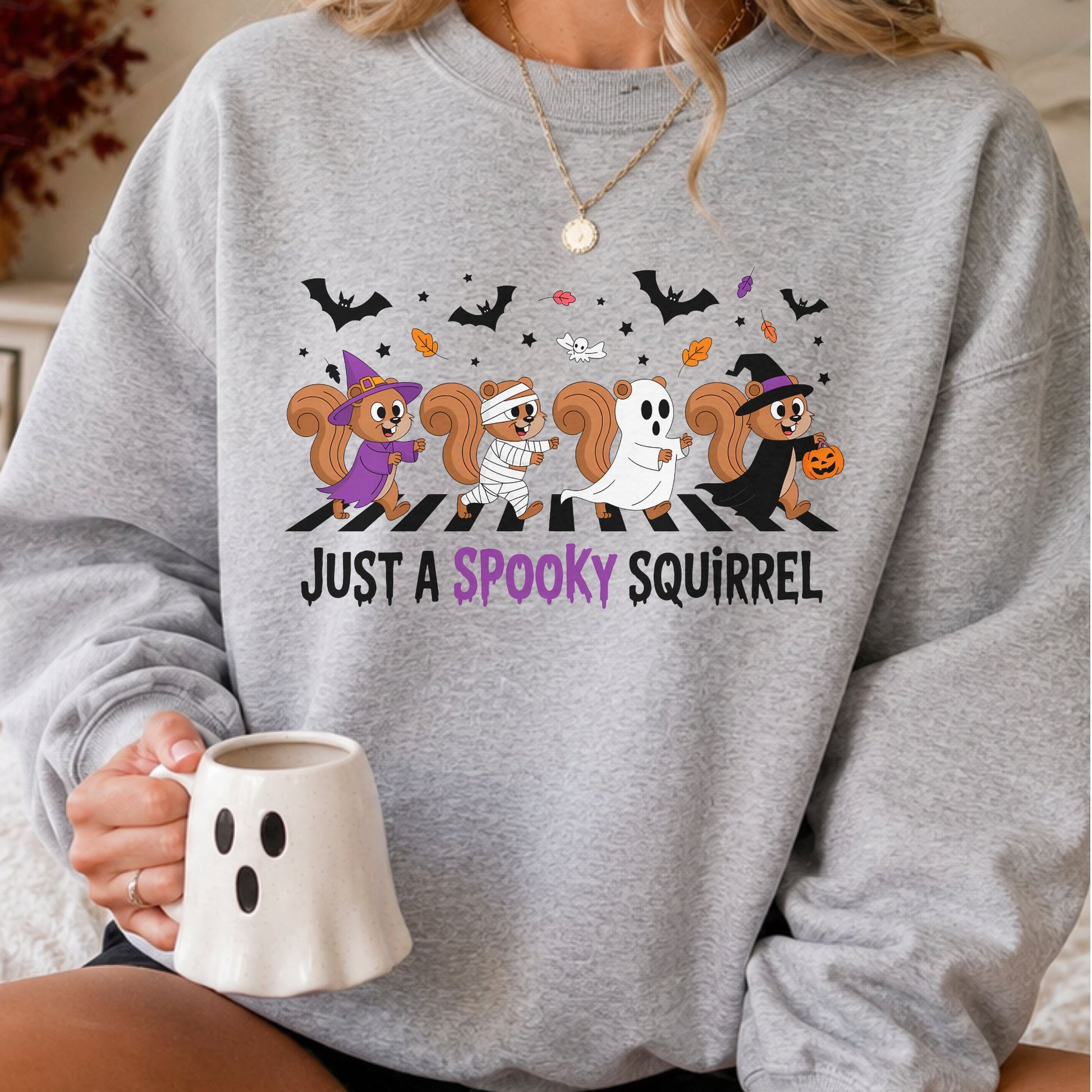 Just a Spooky Squirrel Halloween DTF transfer featuring four playful squirrels dressed as a purple witch, mummy, sheet ghost and black witch, walking across a striped path beneath flying bats, colorful fall leaves and stars on a gray sweatshirt mockup.
