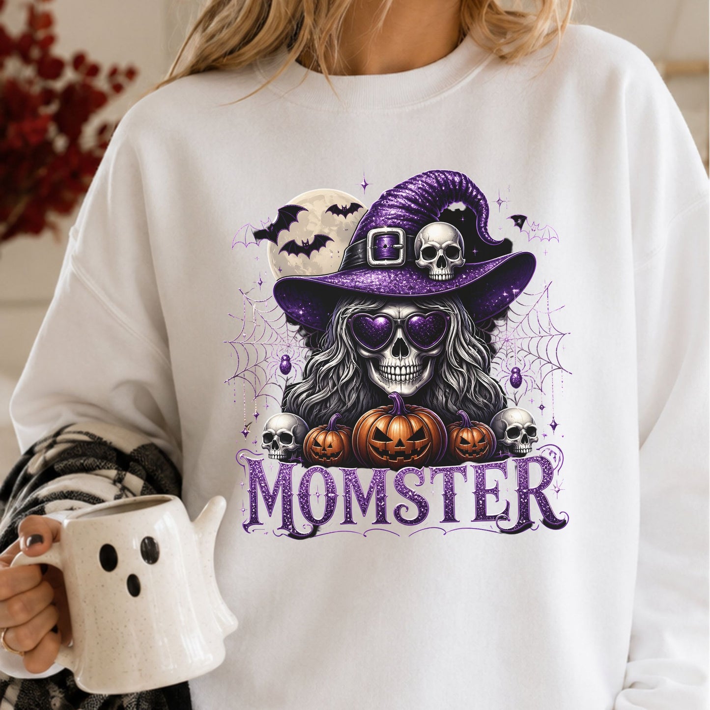 Momster Halloween DTF transfer featuring a glamorous witch skull wearing purple heart-shaped sunglasses and a glittery witch hat, surrounded by jack-o-lantern pumpkins, skulls, bats, spiderwebs and sparkling purple accents on a white sweatshirt mockup.