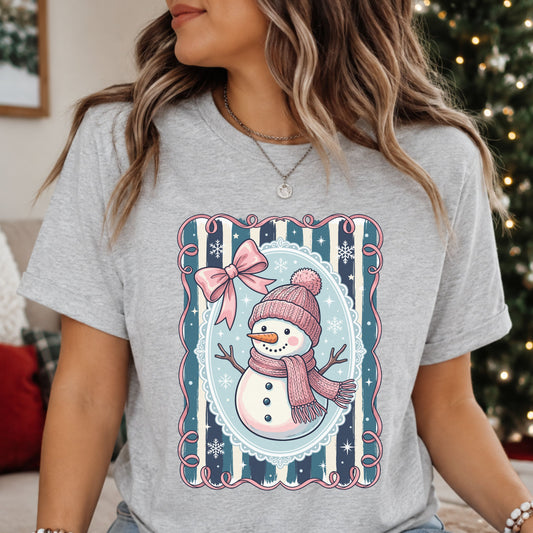 Snowman wearing a pink hat and scarf with a faux knit pattern, surrounded by a pale blue oval frame, navy and teal stripes, a pink bow and white snowflakes.