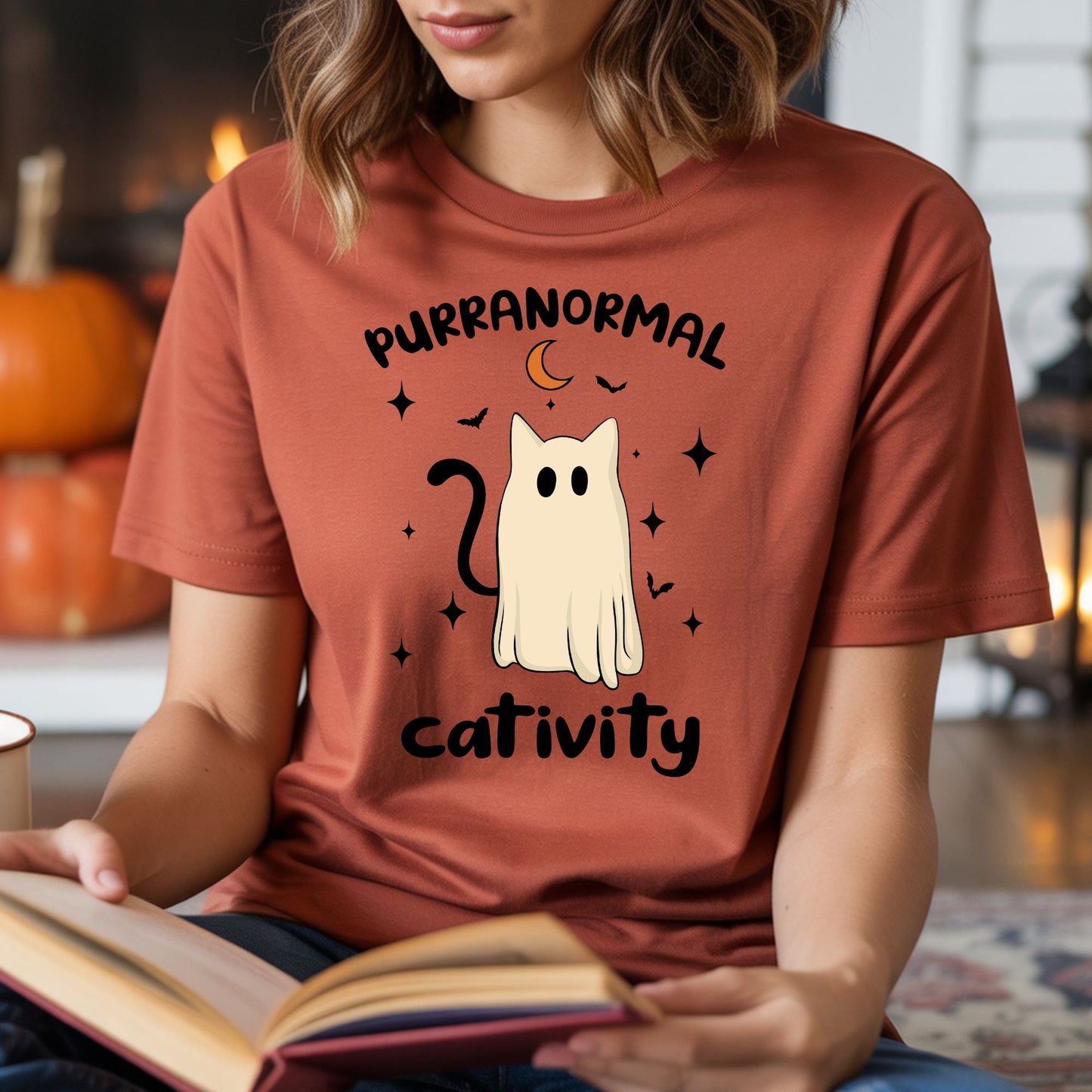 Rust-colored T-shirt mockup featuring a ghost cat design with the phrase Purranormal Cativity, black stars, bats, and a crescent moon in a cute Halloween style.