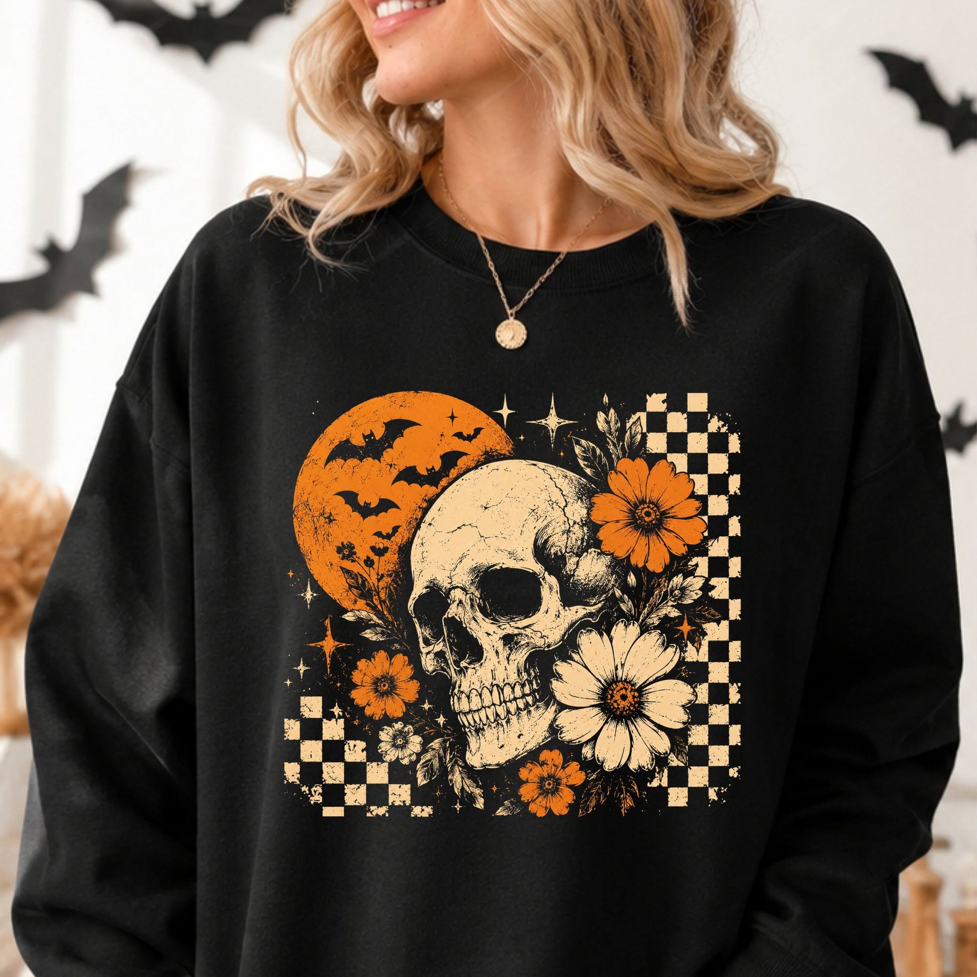Retro floral skull Halloween DTF transfer featuring a detailed cream-colored skull surrounded by orange and ivory flowers, leafy accents, an orange full moon, flying bats, sparkling stars and distressed black-and-cream checkerboard details on a black sweatshirt mockup.