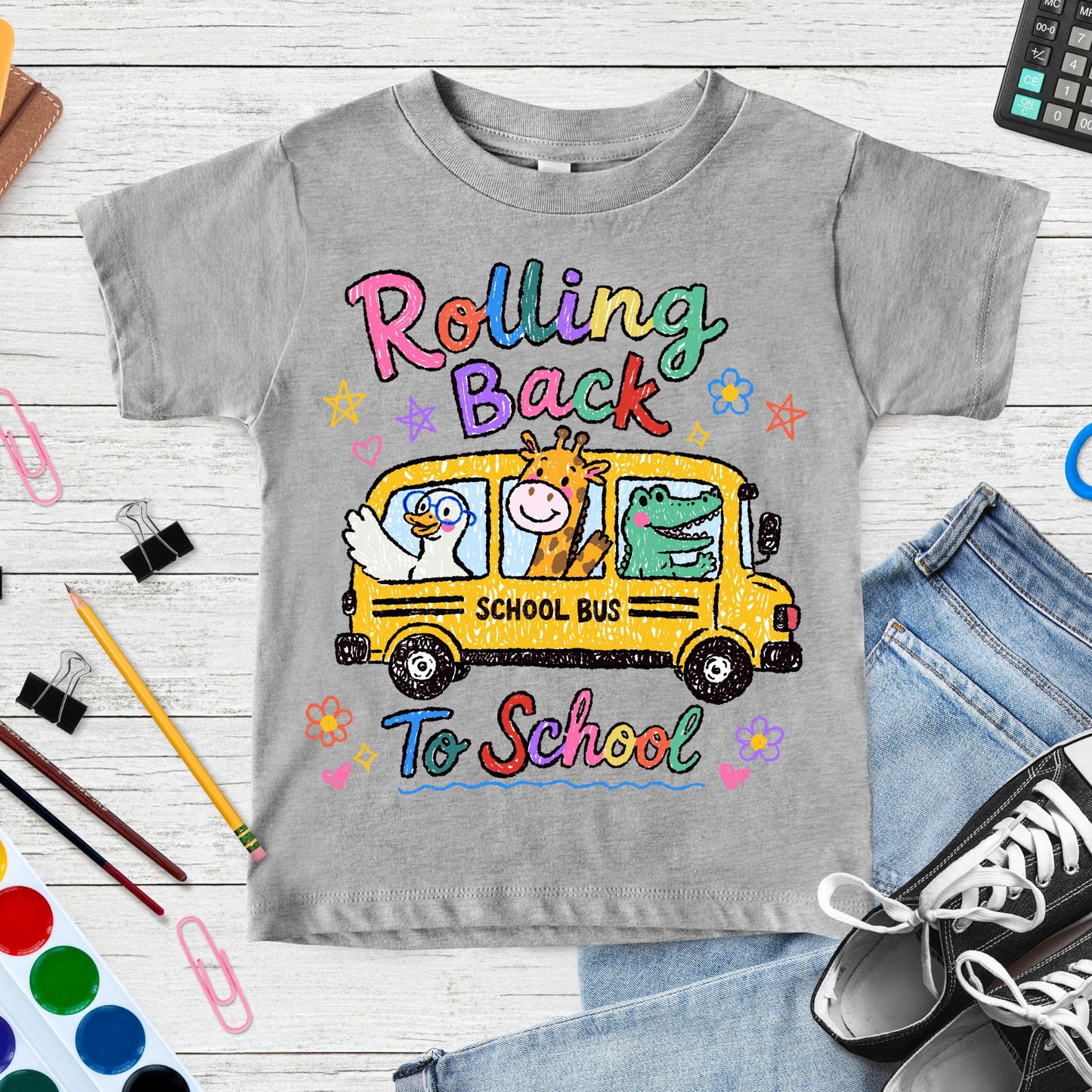 Rolling Back to School design featuring a yellow school bus with a goose, giraffe, and alligator riding inside, surrounded by colorful flowers, stars, hearts, and playful crayon-style lettering.
