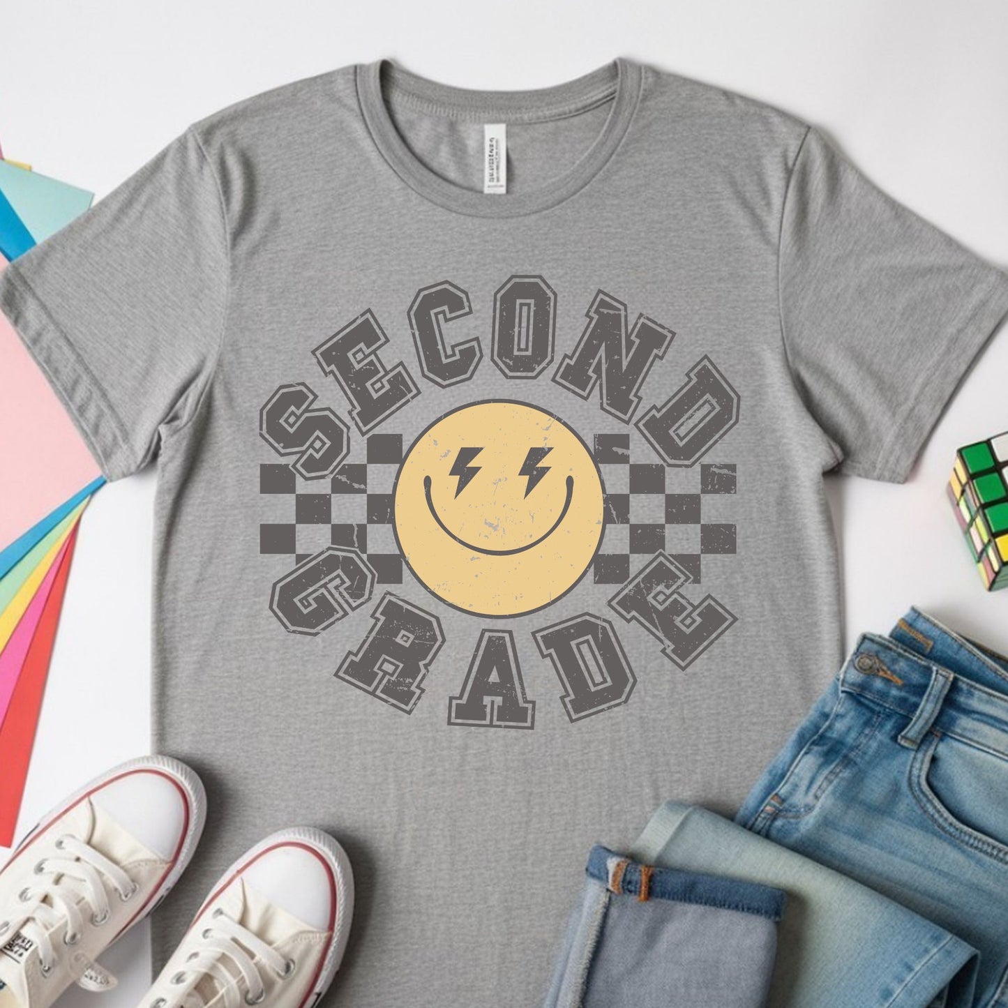 Second Grade back-to-school design featuring distressed collegiate lettering, a yellow smiley face with lightning-bolt eyes, and bold black checkerboard accents for kids shirts.