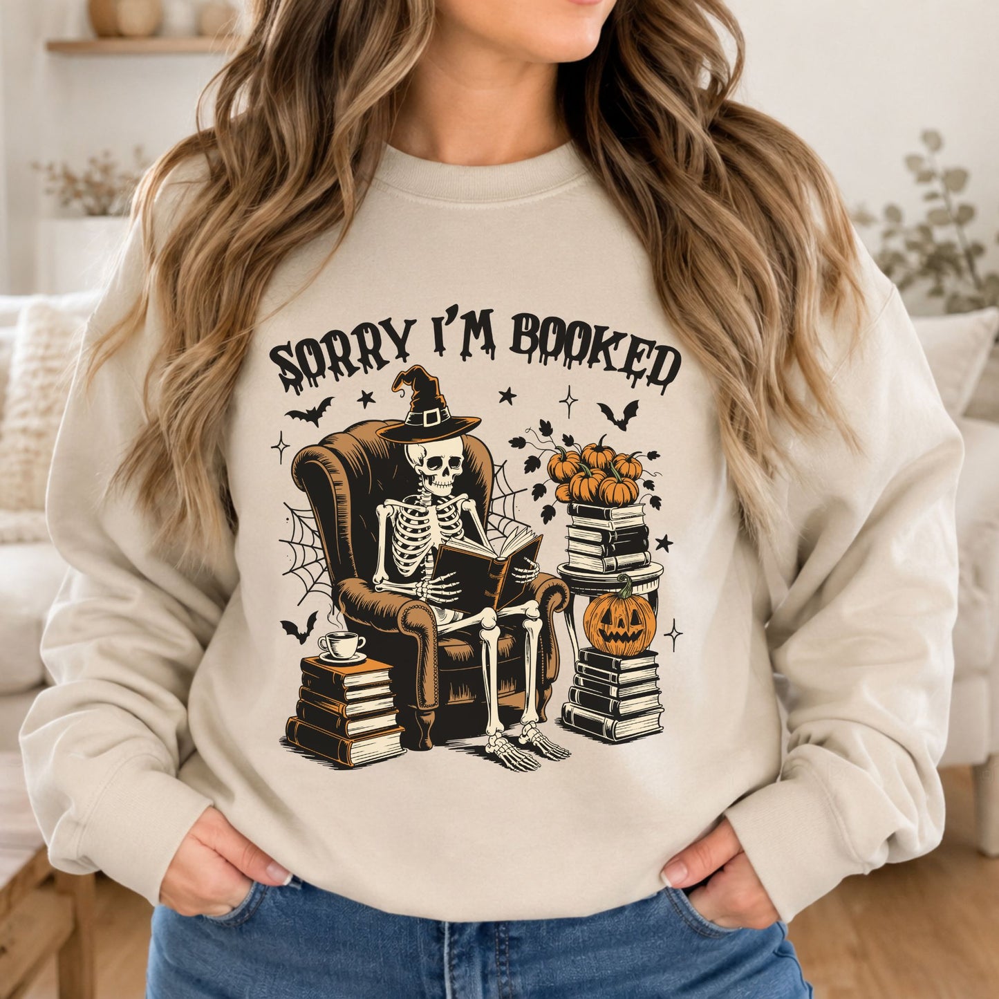 Cream sweatshirt mockup featuring a spooky skeleton reading a book in a chair with pumpkins, stacked books, and the phrase Sorry I’m Booked.
