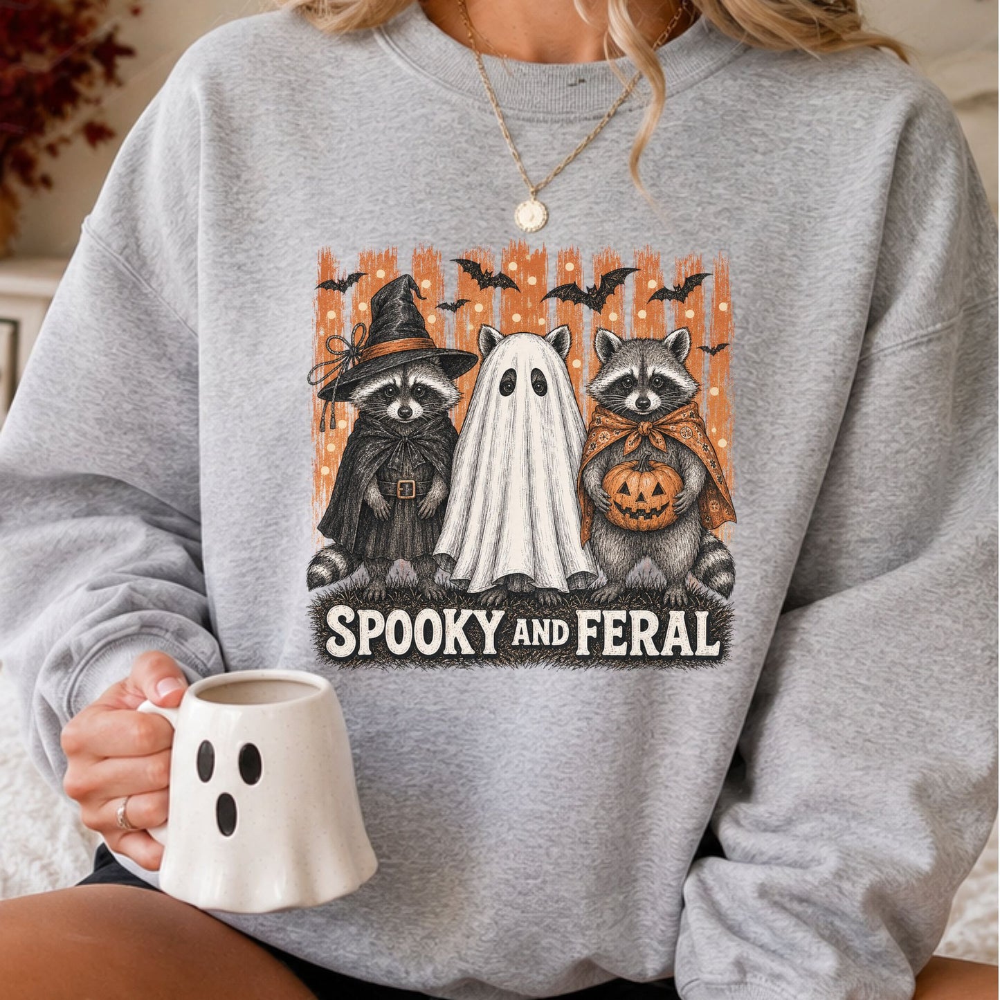 Spooky and Feral Halloween DTF transfer featuring two raccoons dressed as a witch and pumpkin carrier beside a sheet ghost, with flying bats and a distressed orange background on a gray sweatshirt mockup.