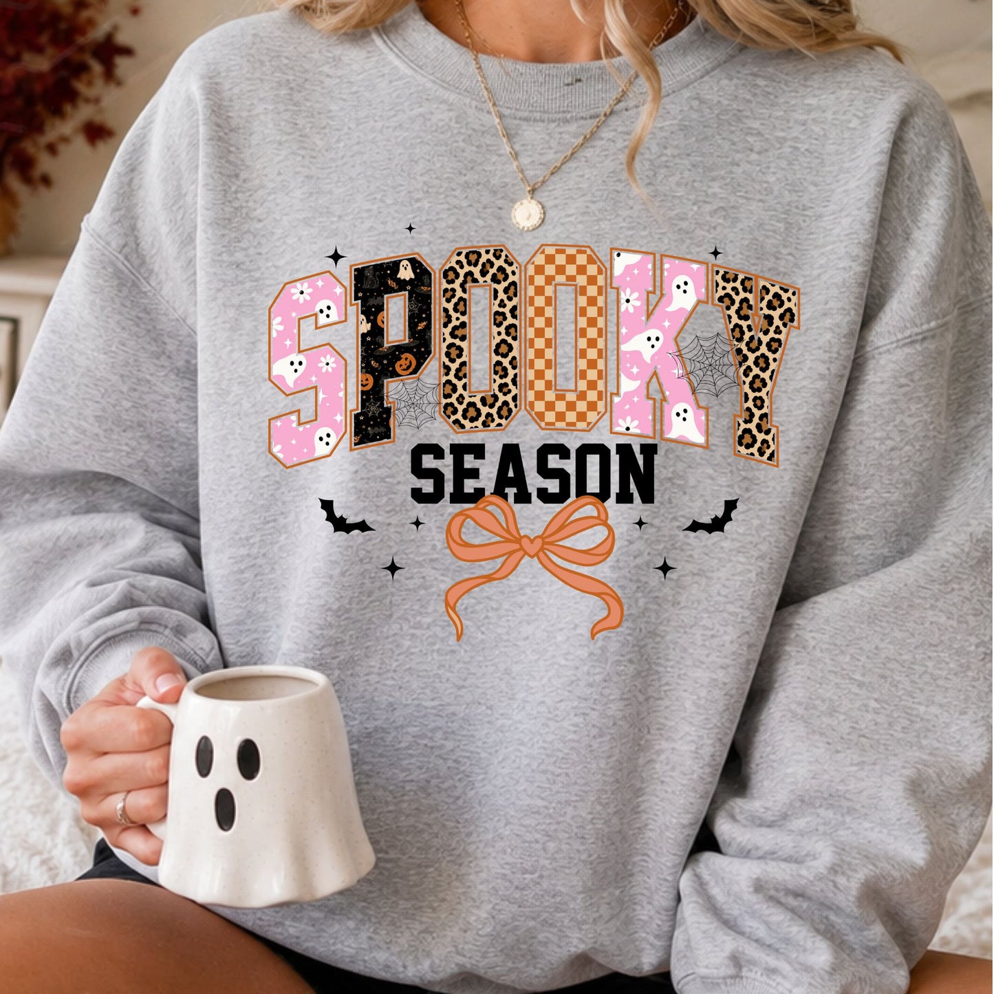 Spooky Season Halloween DTF transfer featuring oversized patterned lettering with pink ghosts, pumpkins, leopard print, orange checkerboard and spiderweb details, paired with an orange coquette bow, bats and sparkling accents on a gray sweatshirt mockup.