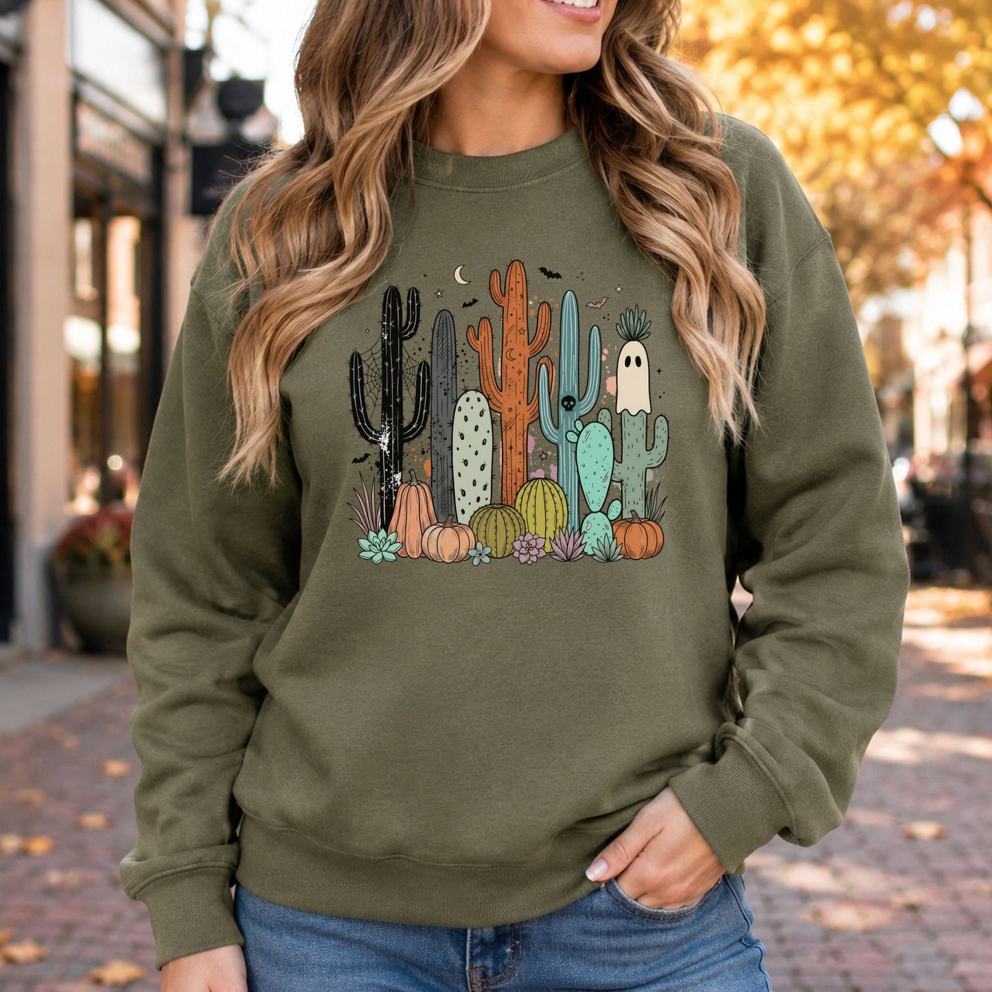 Spooky desert Halloween DTF transfer featuring colorful black, orange, sage green and turquoise cacti surrounded by pumpkins, succulents and desert plants, with a cute white ghost, crescent moon, bats and tiny stars on an olive green sweatshirt mockup.