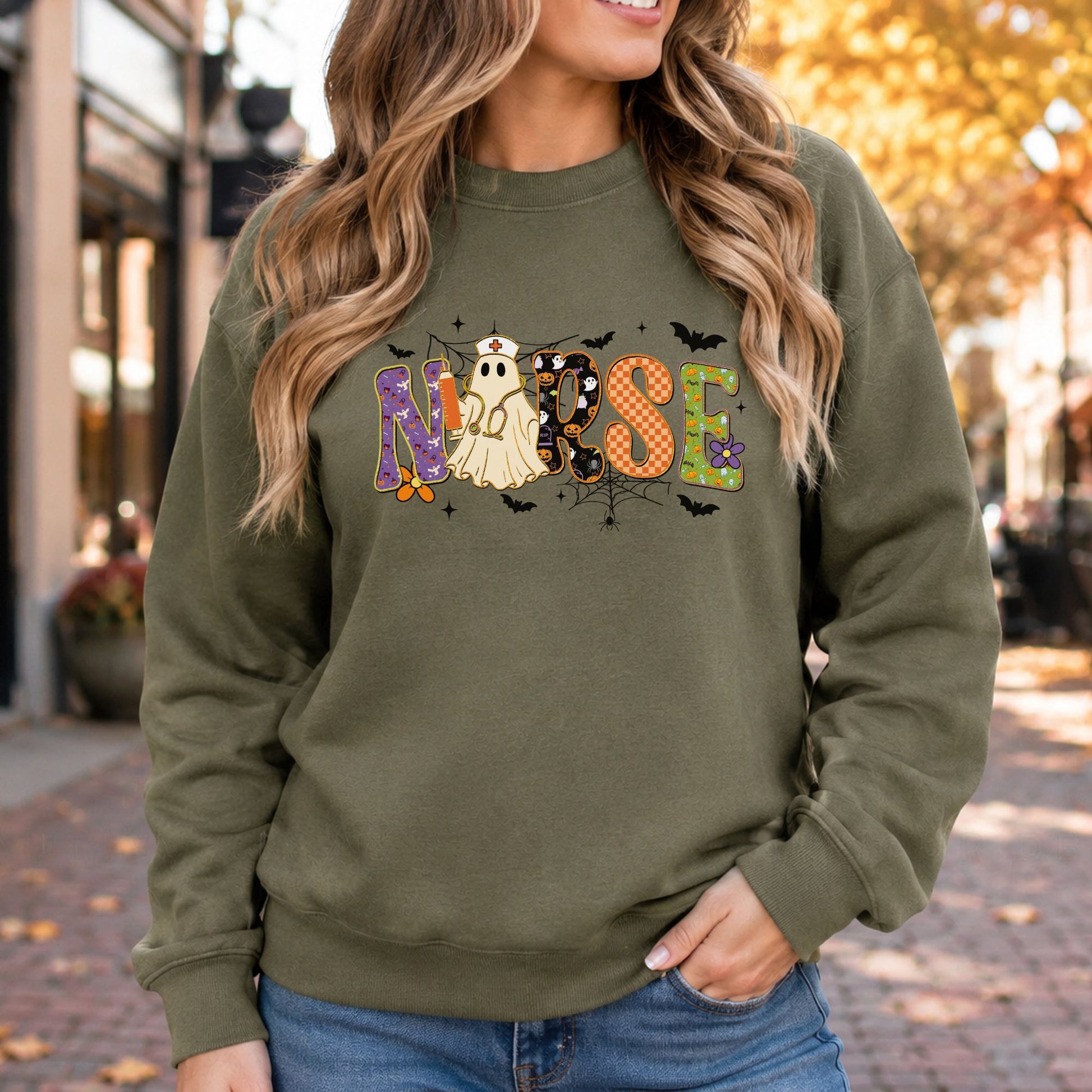 Halloween Nurse DTF transfer featuring colorful patterned NURSE lettering with a sheet ghost dressed as a nurse, holding a syringe and wearing a stethoscope, surrounded by bats, spiderwebs, stars, pumpkins and floral accents on an olive green sweatshirt mockup.