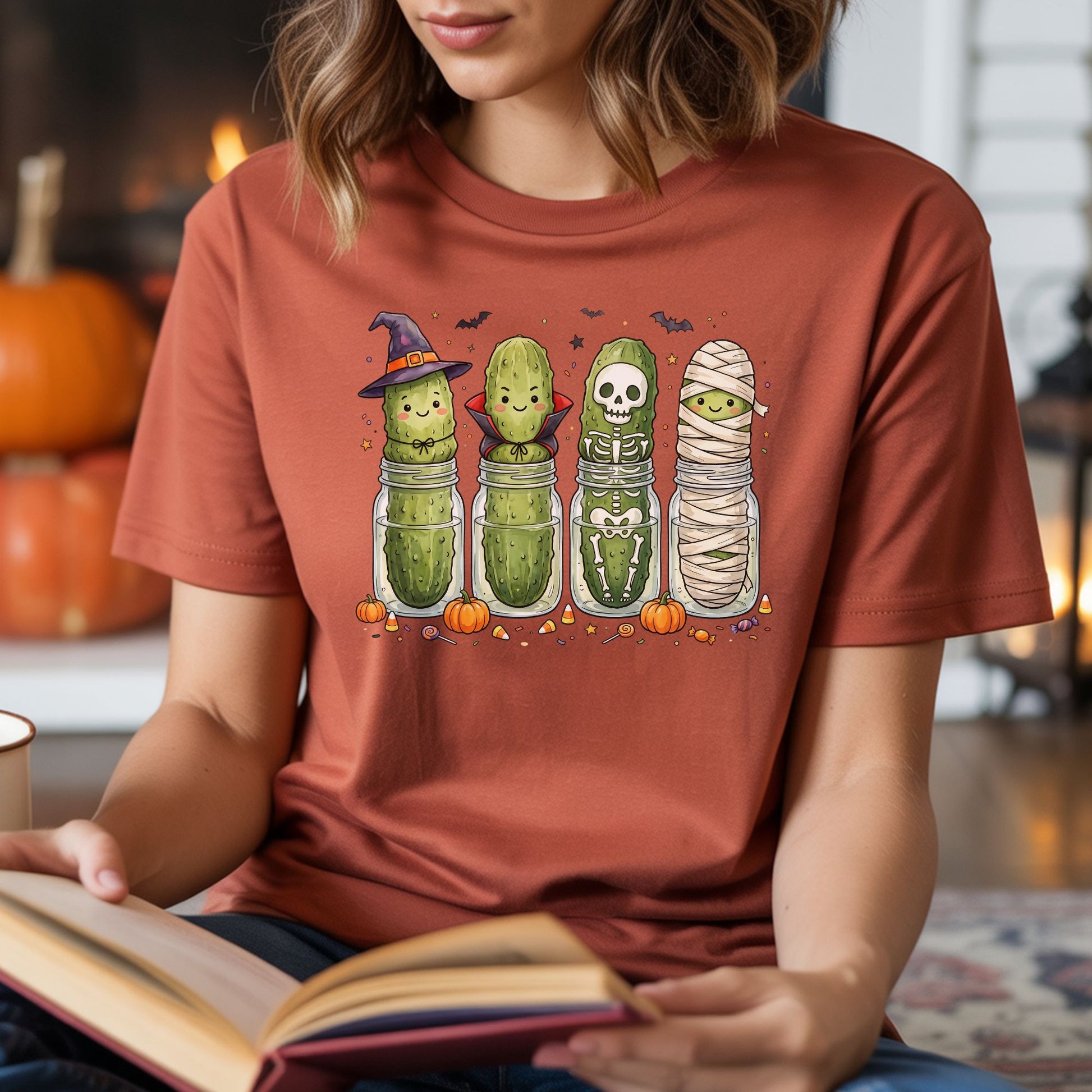 Rust-colored T-shirt mockup featuring four cute Halloween pickles dressed as a witch, vampire, skeleton, and mummy with pumpkins, bats, and candy.