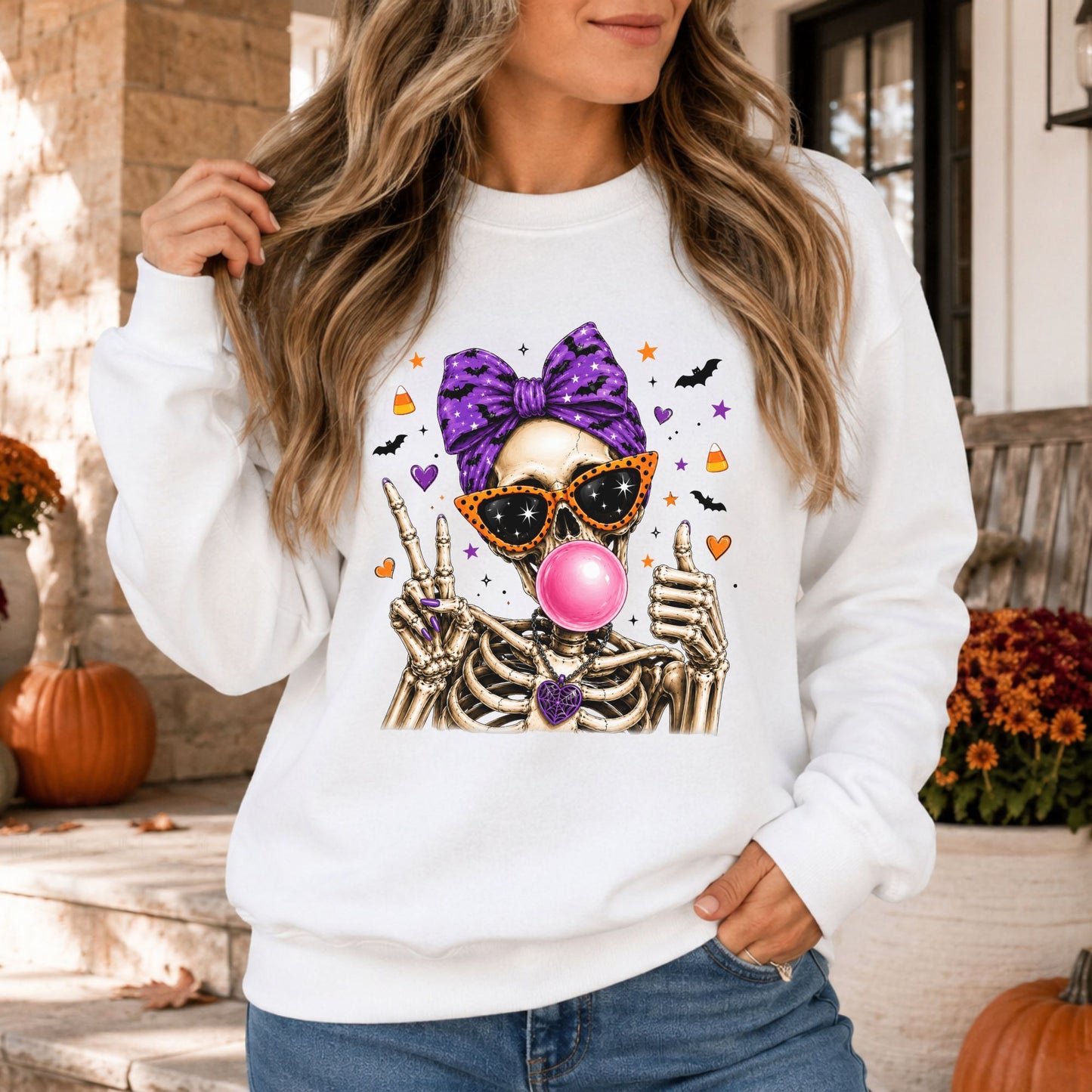 White sweatshirt mockup featuring a spooky skeleton blowing pink bubblegum while wearing orange sunglasses and a purple bow with Halloween bats and candy corn.