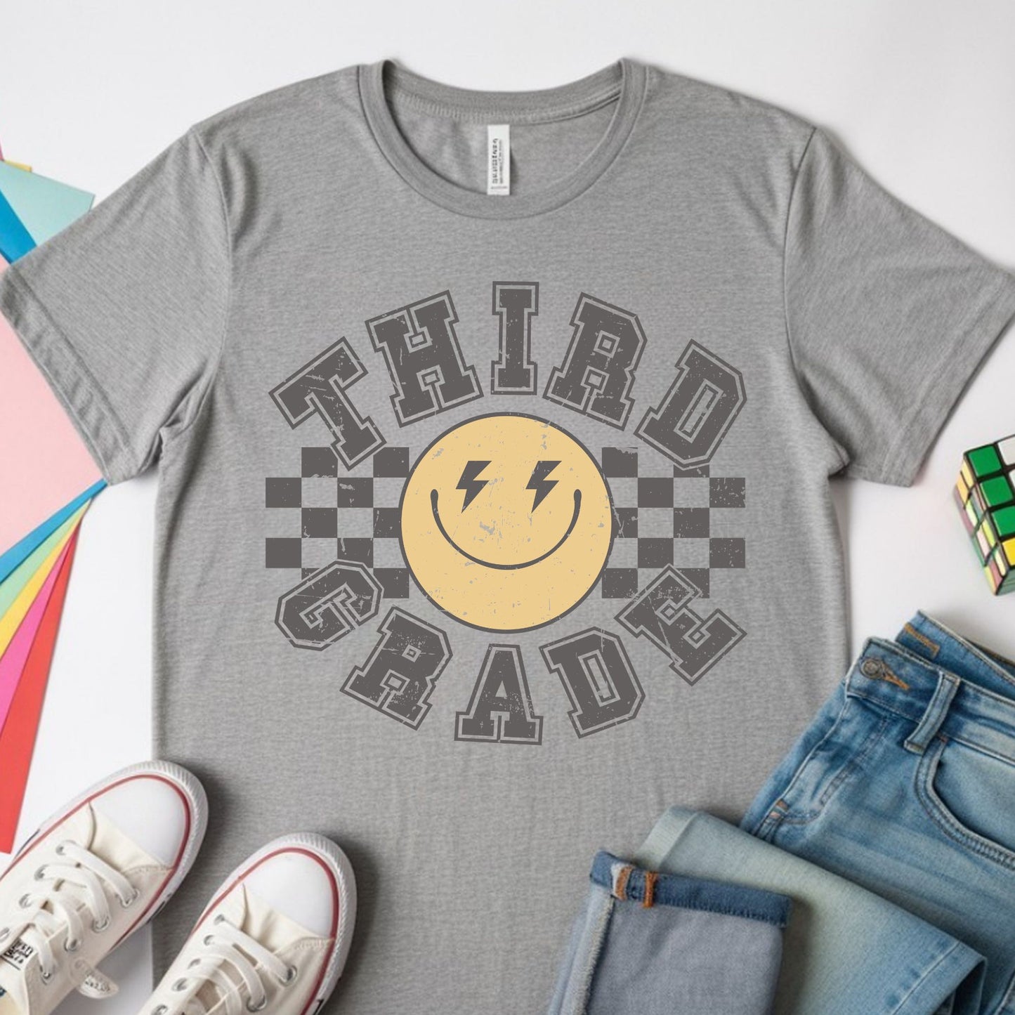 Third Grade back-to-school design featuring distressed collegiate lettering, a yellow smiley face with lightning-bolt eyes, and bold black checkerboard accents for kids shirts.