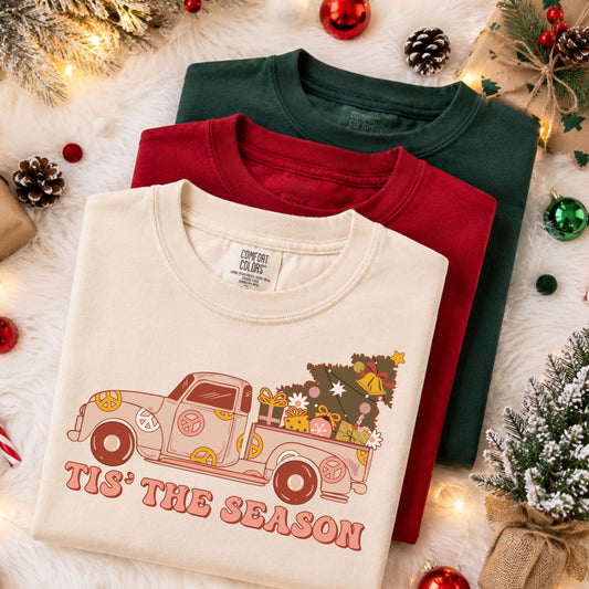 Tis’ the Season beneath a peach vintage pickup decorated with pink, mustard and cream peace symbols, carrying a Christmas tree, gifts, ornaments, daisies and bells.