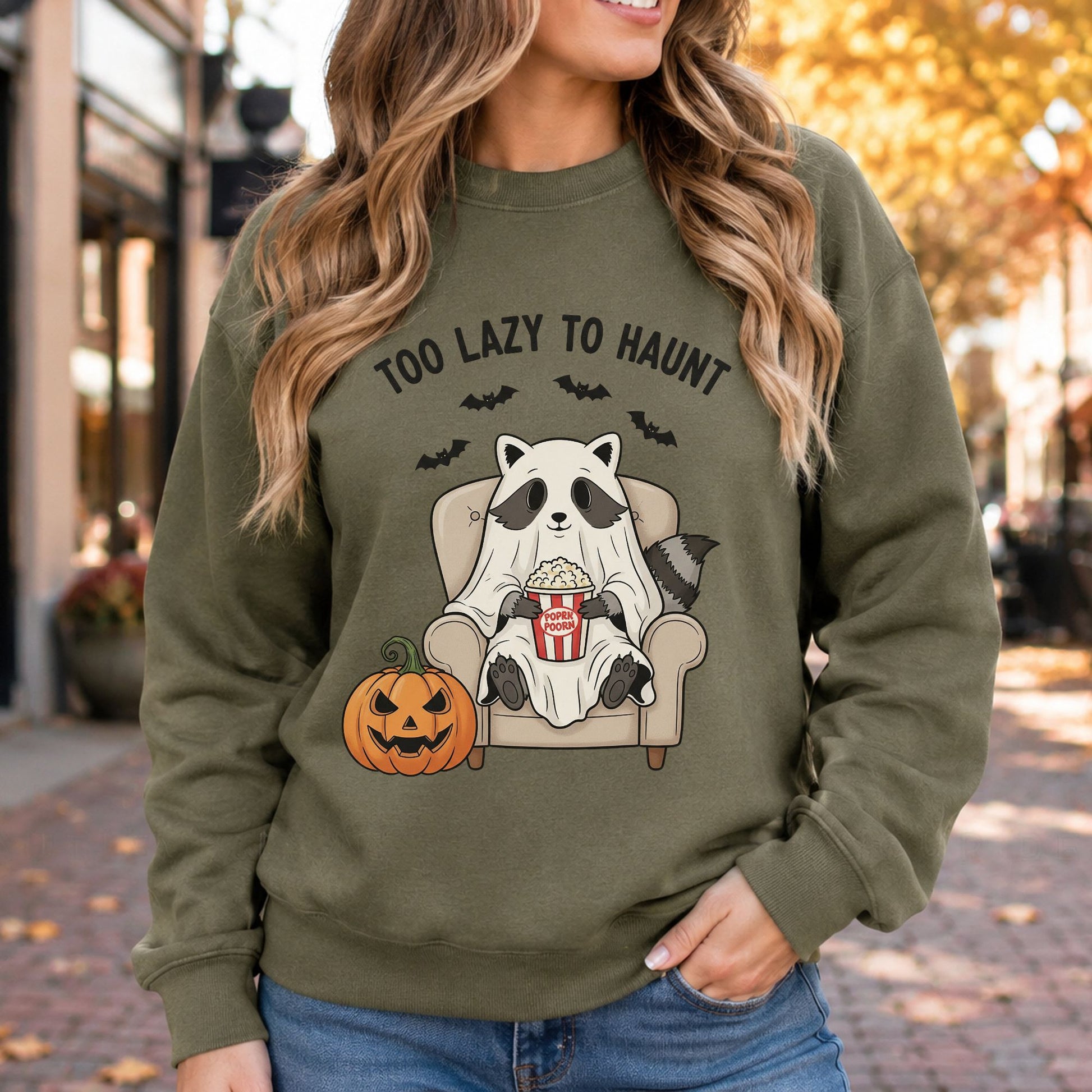 Too Lazy to Haunt Halloween DTF transfer featuring a cute raccoon dressed as a sheet ghost, relaxing in an armchair with a tub of popcorn beside a jack-o’-lantern, with flying bats above on an olive green sweatshirt mockup.