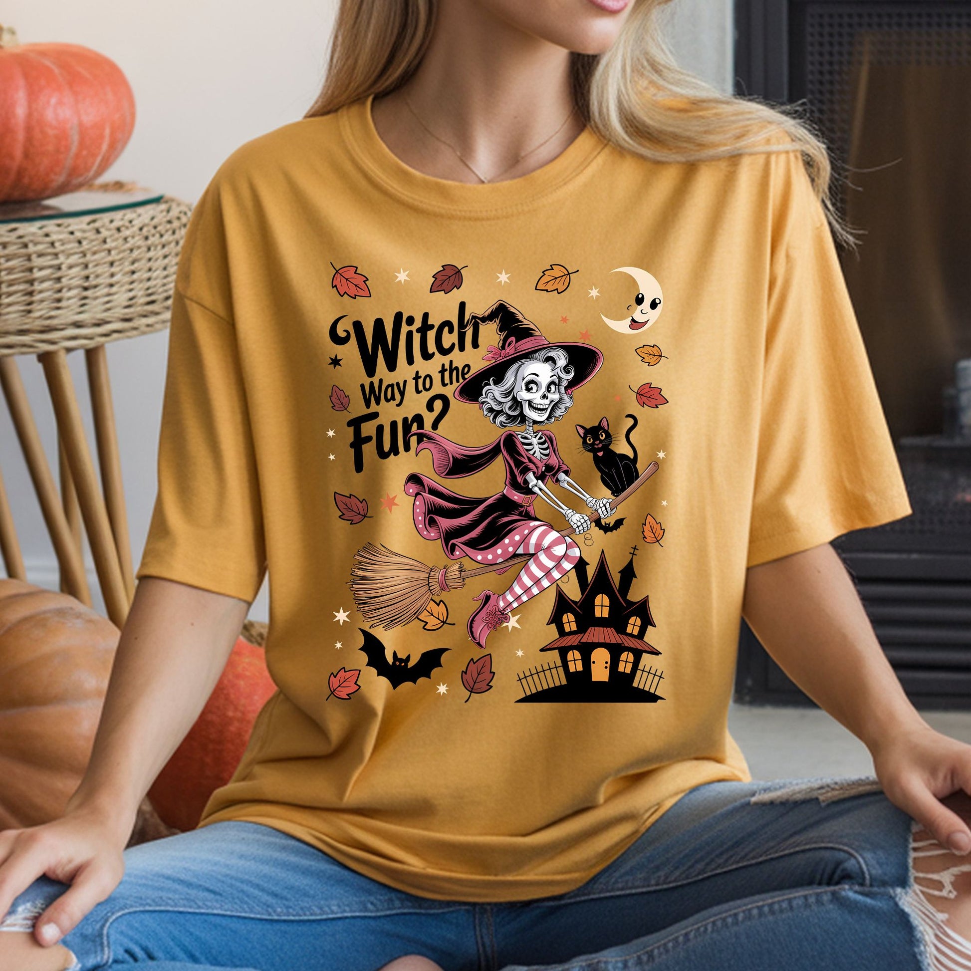 Witch Way to the Fun Halloween DTF transfer featuring a cheerful skeleton witch flying on a broom with a black cat, haunted house, smiling crescent moon, bats, stars and colorful fall leaves in pink, orange and black on a mustard yellow T-shirt mockup.
