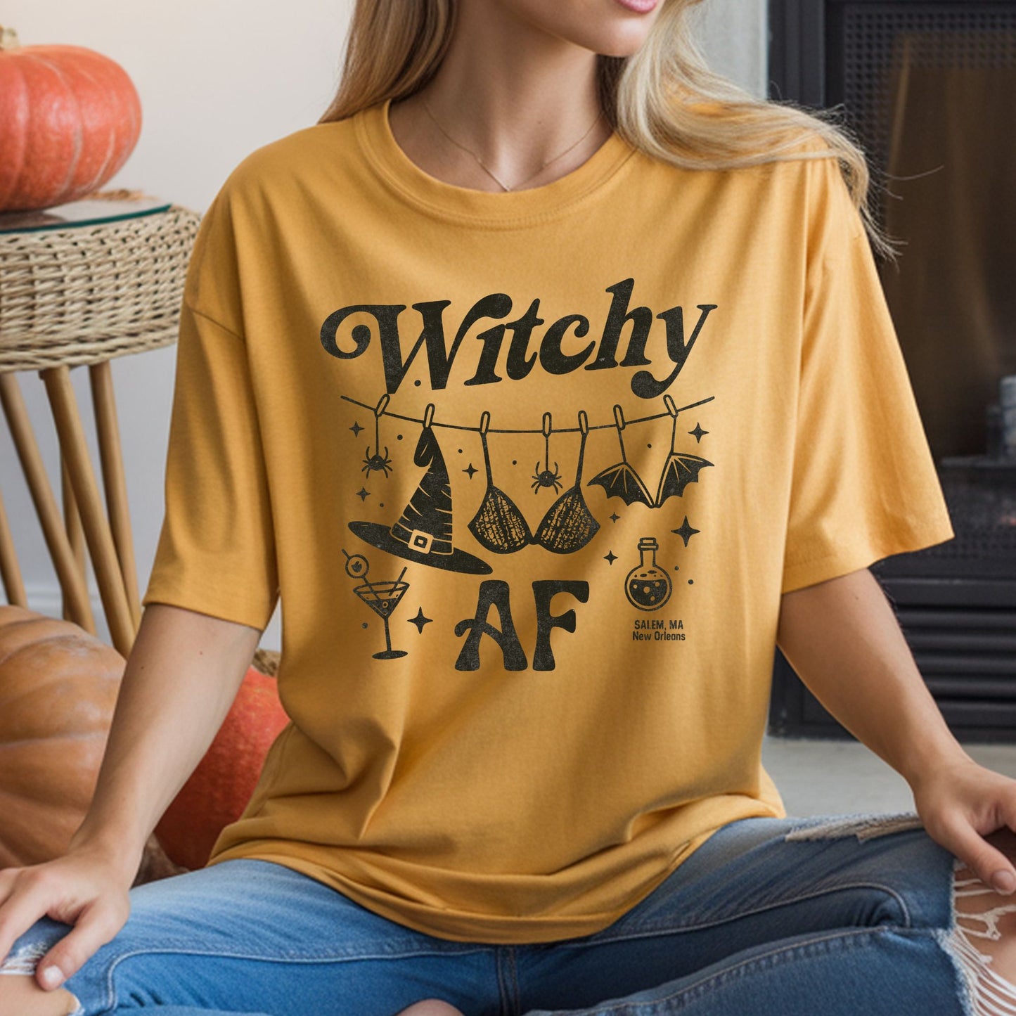 Witchy AF Halloween DTF transfer featuring distressed black lettering, a clothesline with a witch hat, lace bra and bat, plus spiders, stars, a cocktail glass and potion bottle with Salem, MA and New Orleans text on a mustard yellow T-shirt mockup.