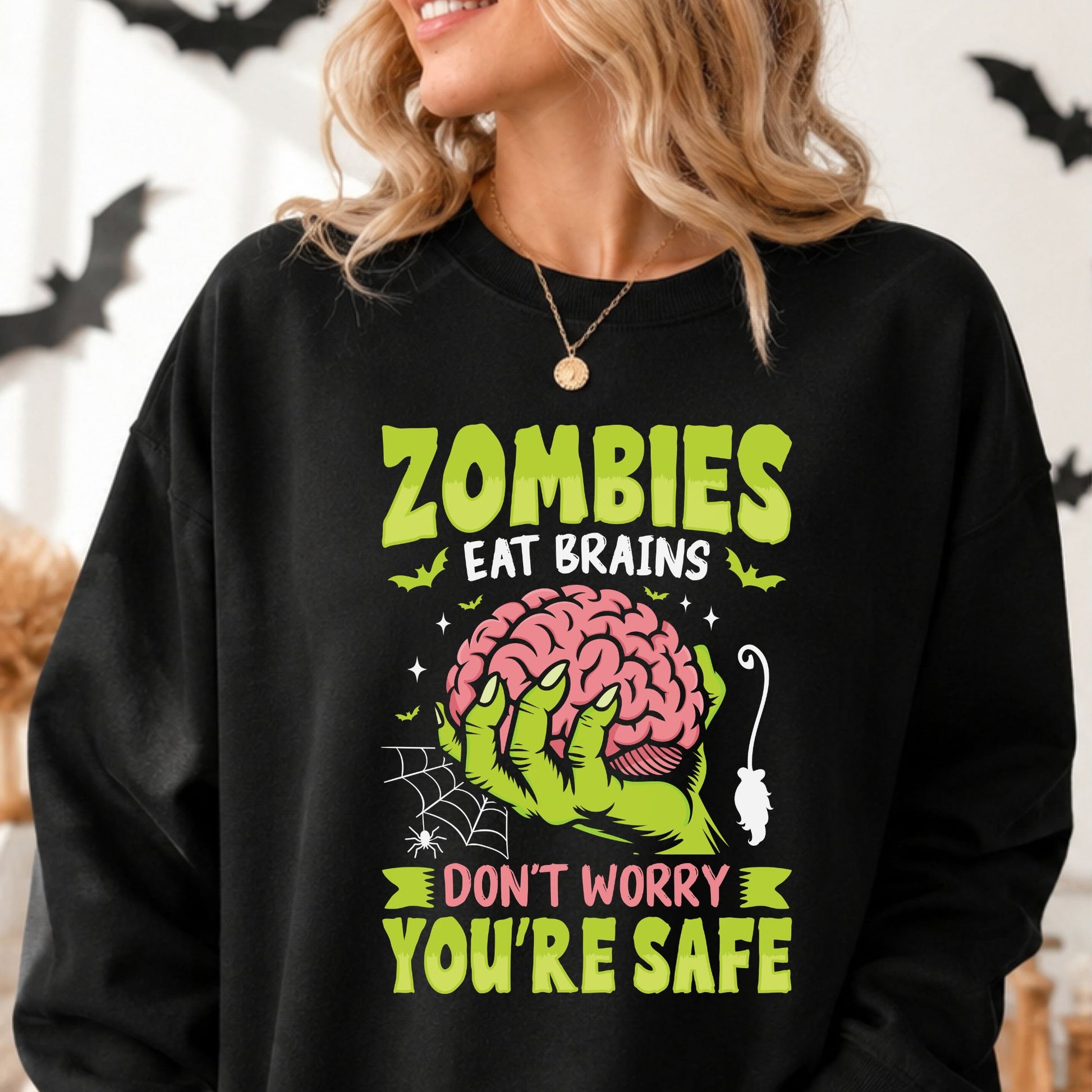 Zombies Eat Brains Don’t Worry You’re Safe Halloween DTF transfer featuring a neon green zombie hand holding a pink brain, surrounded by bats, stars, a spiderweb, spider and dangling mouse on a black sweatshirt mockup.